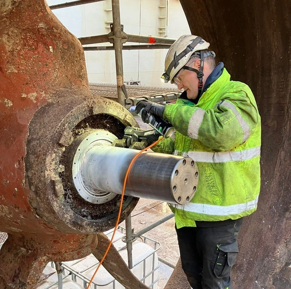 A worker wearing a safety helmet, glasses, and a yellow-green reflective jacket is inspecting or repairing a large metal pipe or machinery component on an industrial site. The worker is holding a spray can and is closely examining the part.