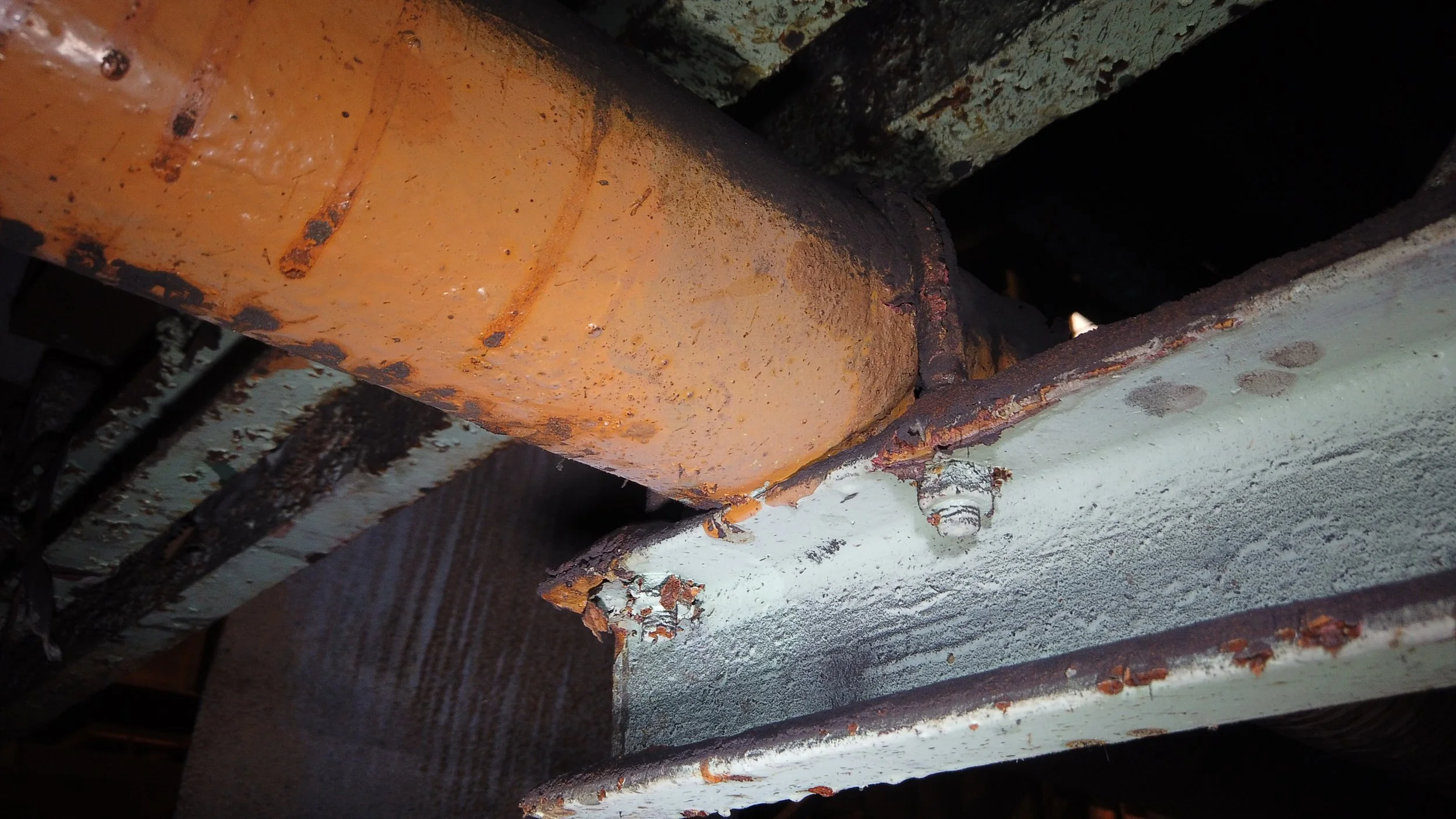 Close-up of a rusty, orange-painted metal pipe connected to a rusty metal surface, with visible rust and corrosion on both.