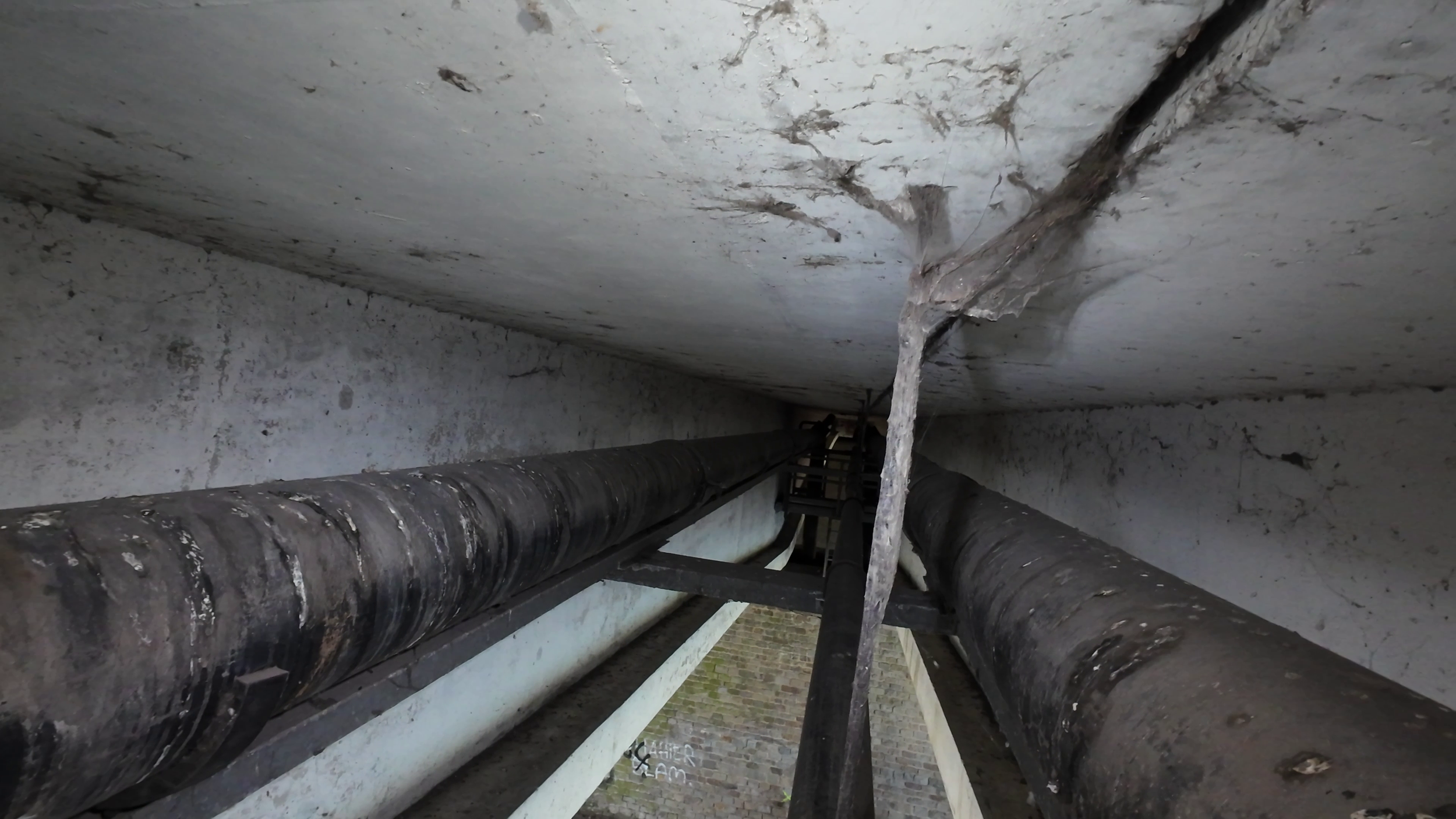 View of the underside of a building with large, black pipes running parallel to each other, with one pipe having peeling paint and corrosion. The ceiling above is concrete with cracks and dirt, and there are supporting beams.