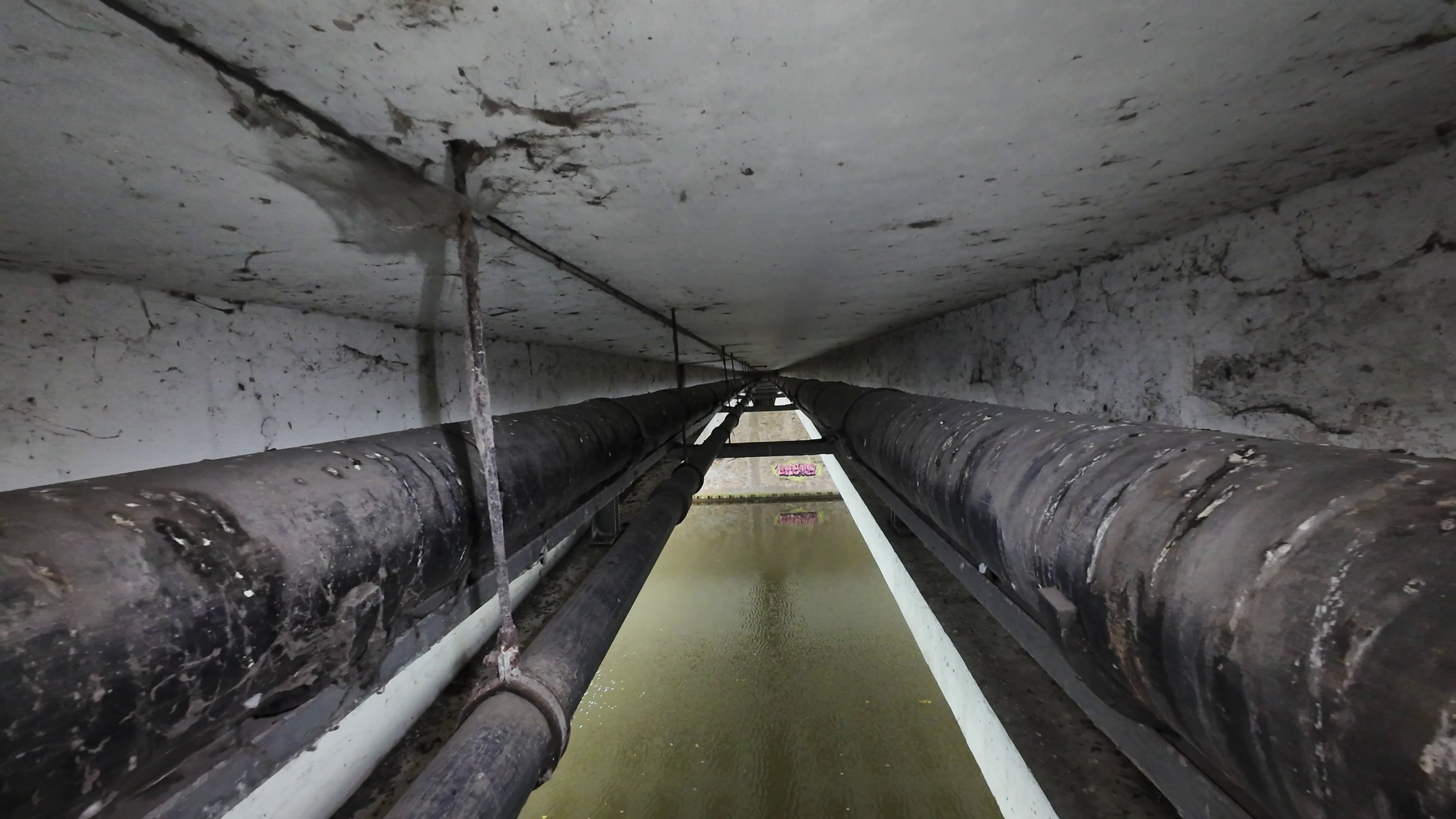 Under a bridge or dam, showing two large black pipes running parallel over water, with a concrete ceiling above and water below.