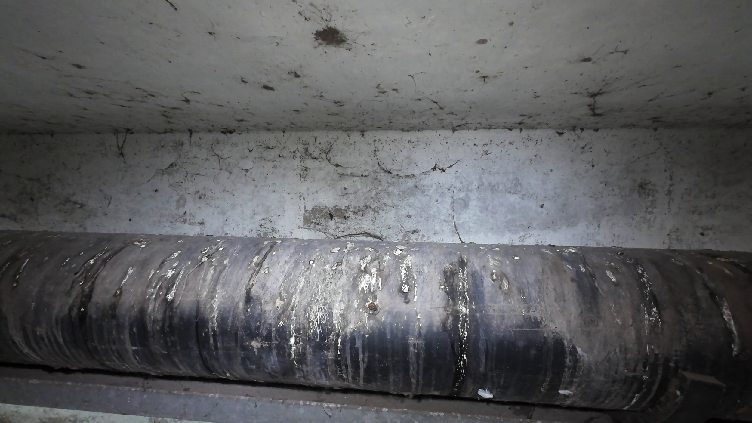 A large, black, industrial pipe with white deposits, mounted horizontally under a ceiling with cracks and stains.