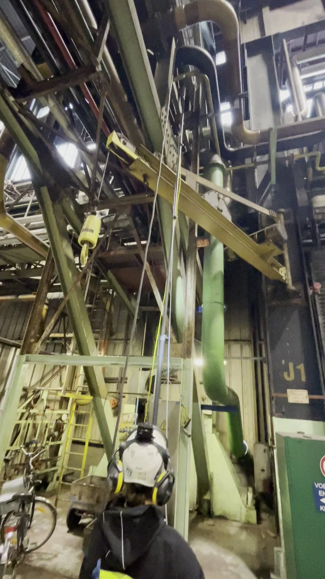 A person with a backpack and yellow and black headphones on, standing in an industrial or mechanical setting, facing equipment and pipes. The photo is taken indoors with metal structures, pipes, and machinery.