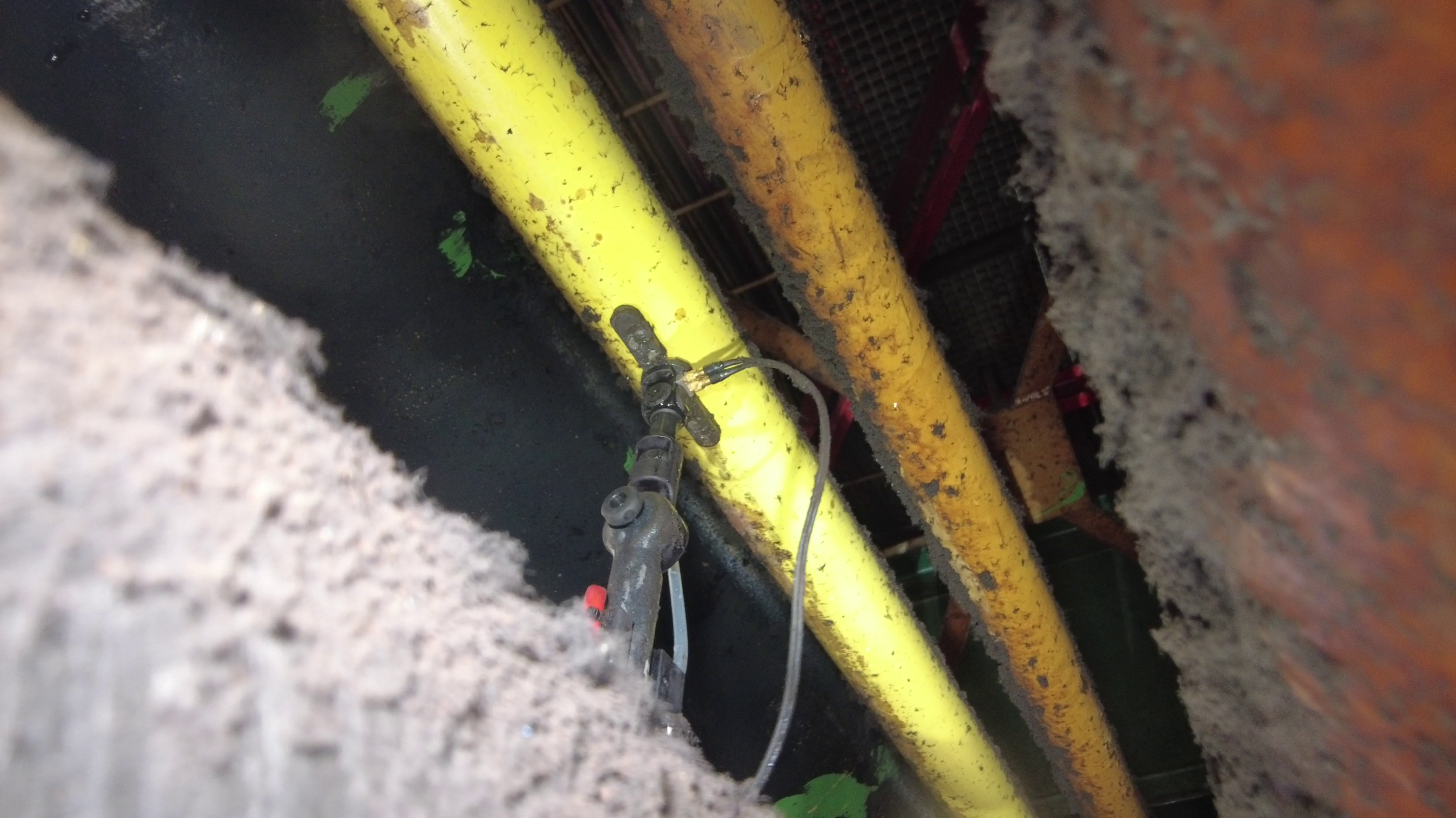 Close-up view of yellow and orange pipes and a valve or connector, seen through a small opening in a wall, with some green foliage visible in the background.