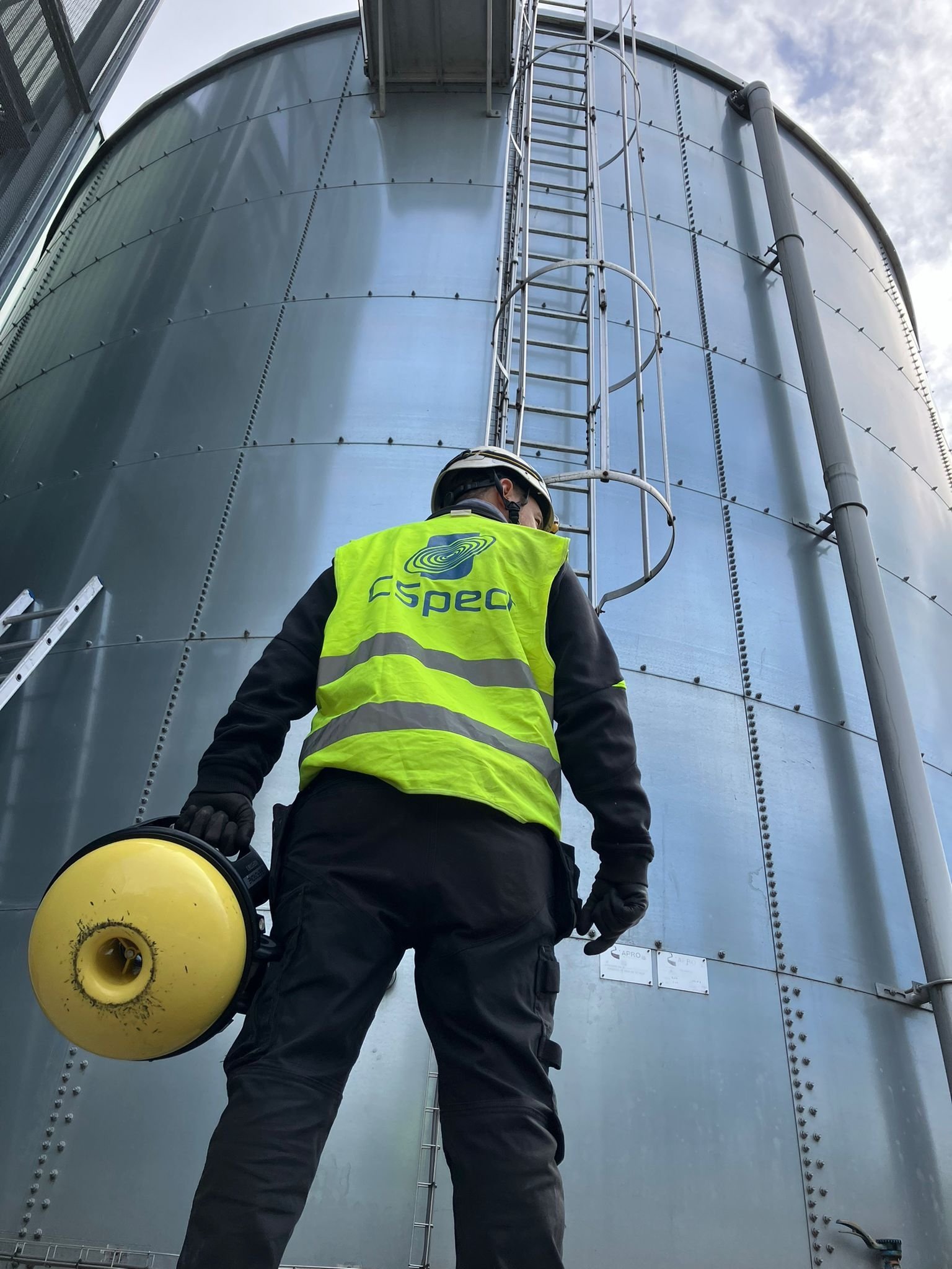 Worker in safety gear holding a beekeeping smoker looking at a large metal storage tank with ladder.