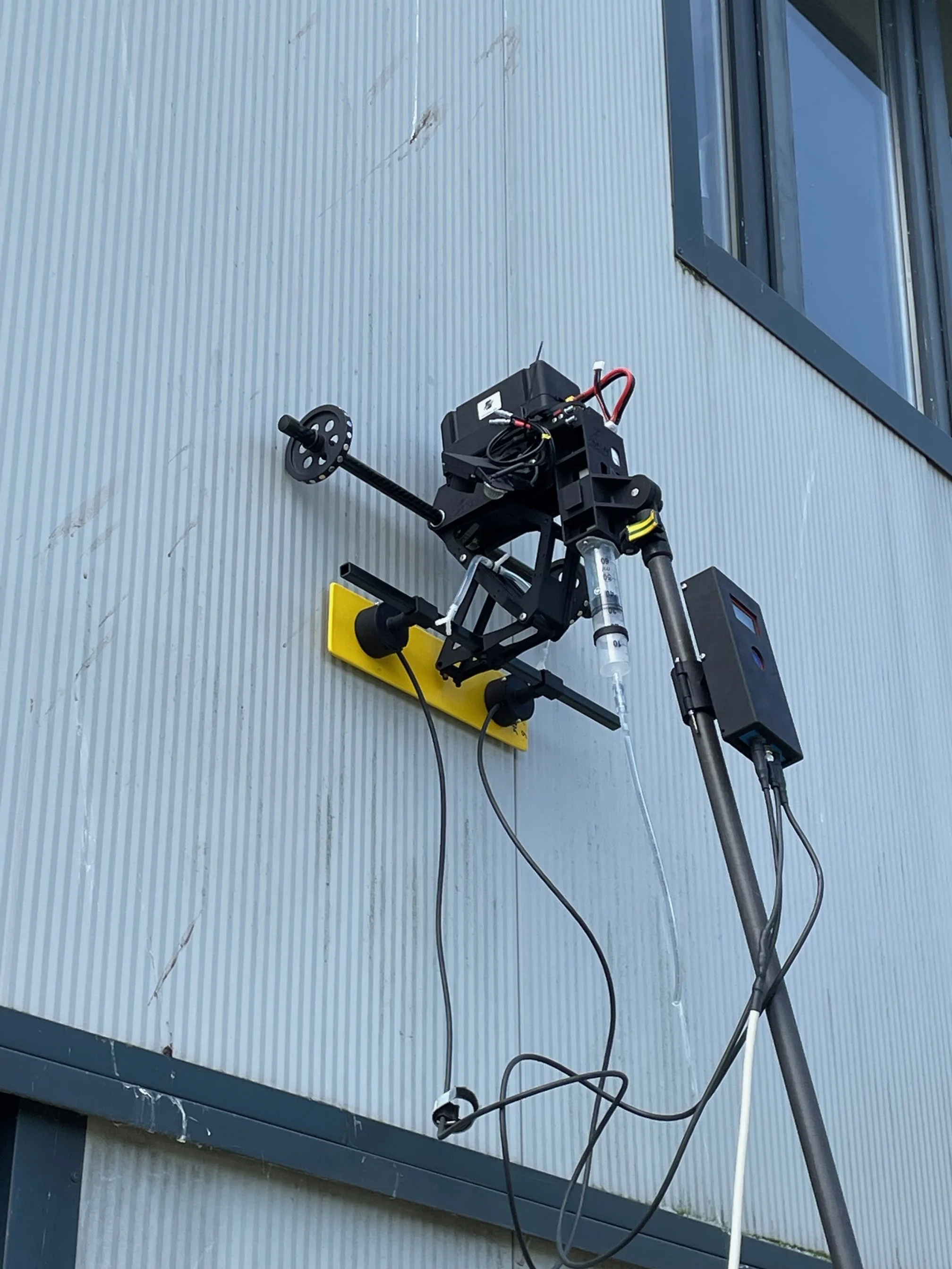 A scientific instrument mounted on a yellow base attached to a gray, corrugated wall outside, with wires connected to various components for research or testing.