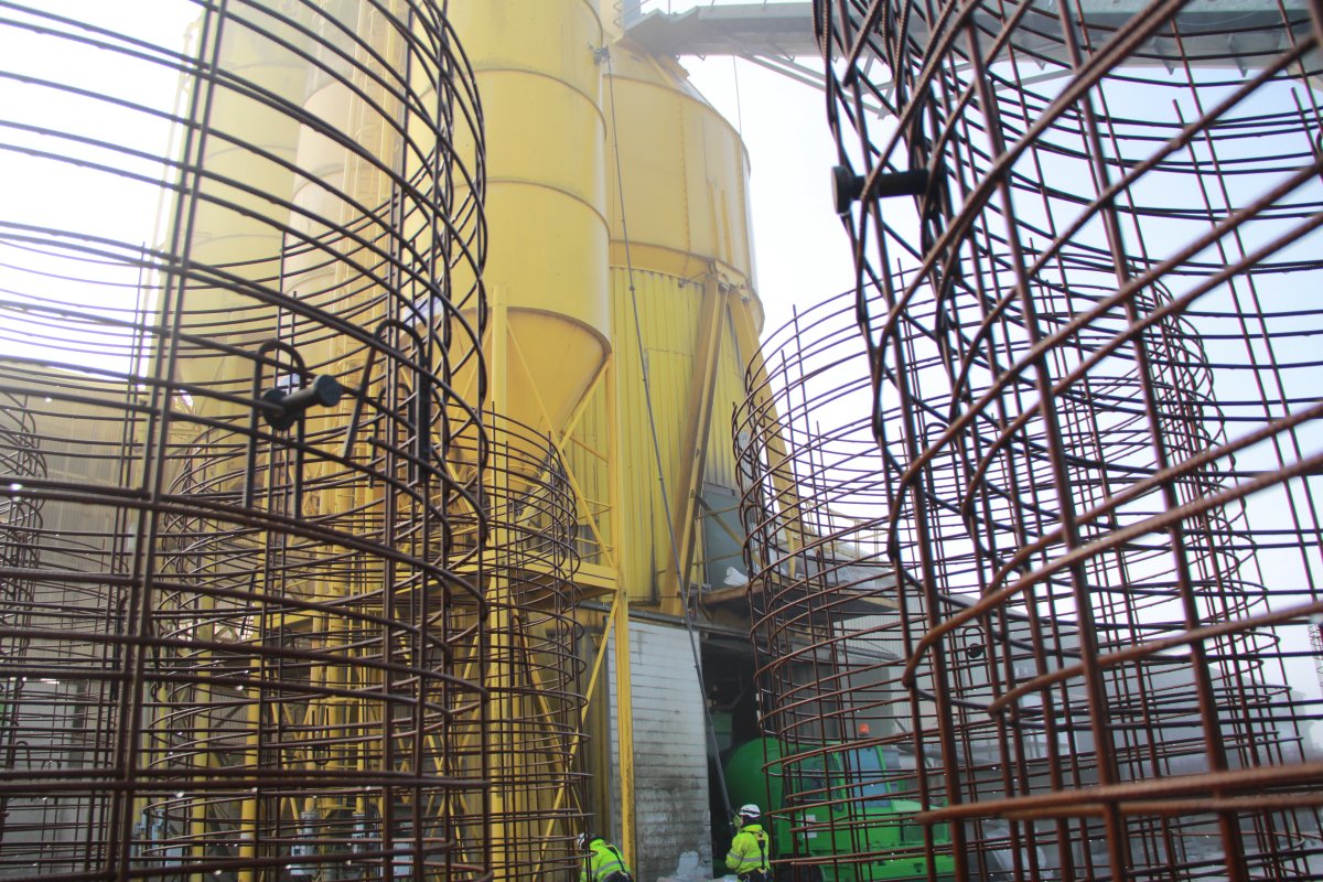 Construction site with large yellow industrial tanks and multiple wire cages in the foreground, with workers wearing safety gear.