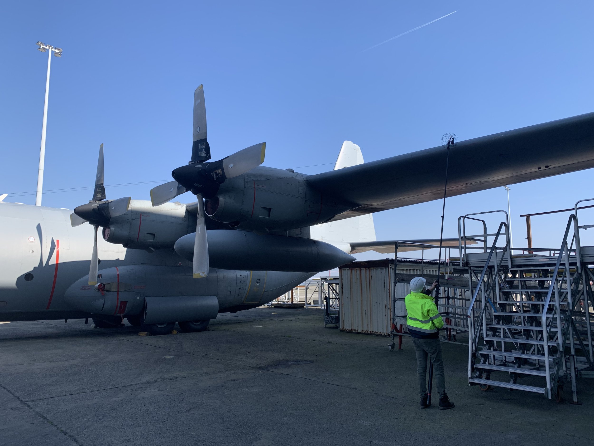 A military aircraft with propellers parked on the tarmac. A person in a high-visibility vest and helmet stands nearby, working near temporary construction or servicing equipment.