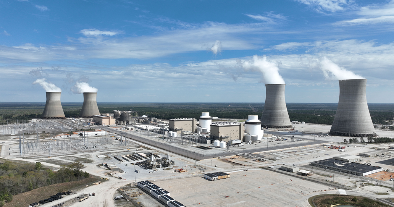 A large power plant with five cooling towers emitting steam, situated in a flat landscape with some small buildings and equipment in the foreground, under a partly cloudy sky.