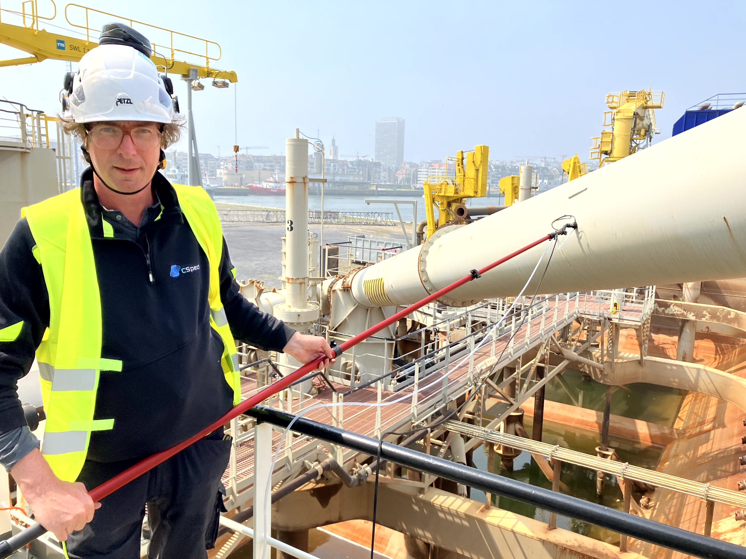 A man wearing a safety helmet, glasses, and a high-visibility vest standing on a ship's deck near large industrial pipes with a city skyline in the background.