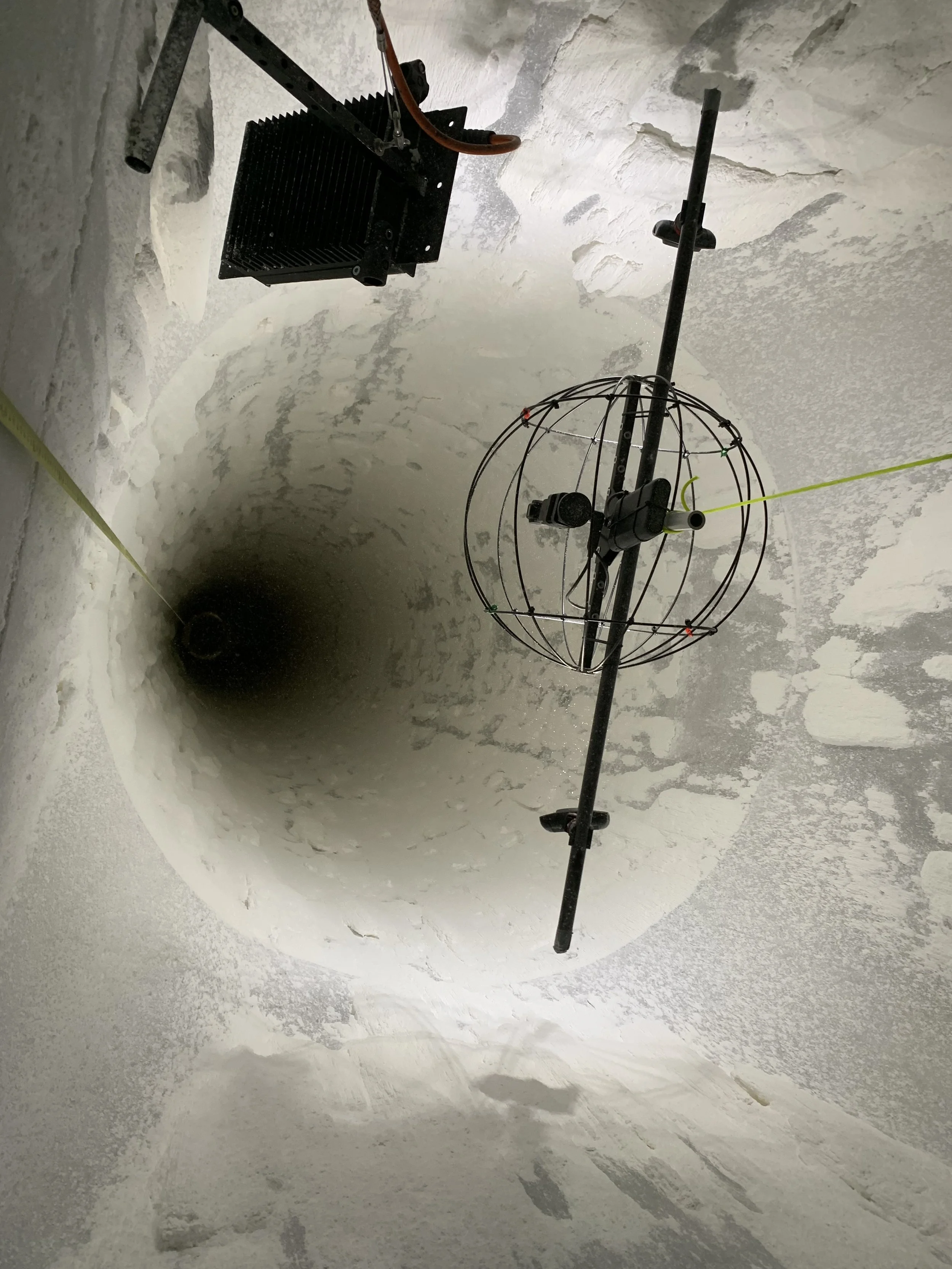 A large vertical hole or shaft in a concrete or rocky surface, with tools and equipment attached, indicating construction or excavation work.