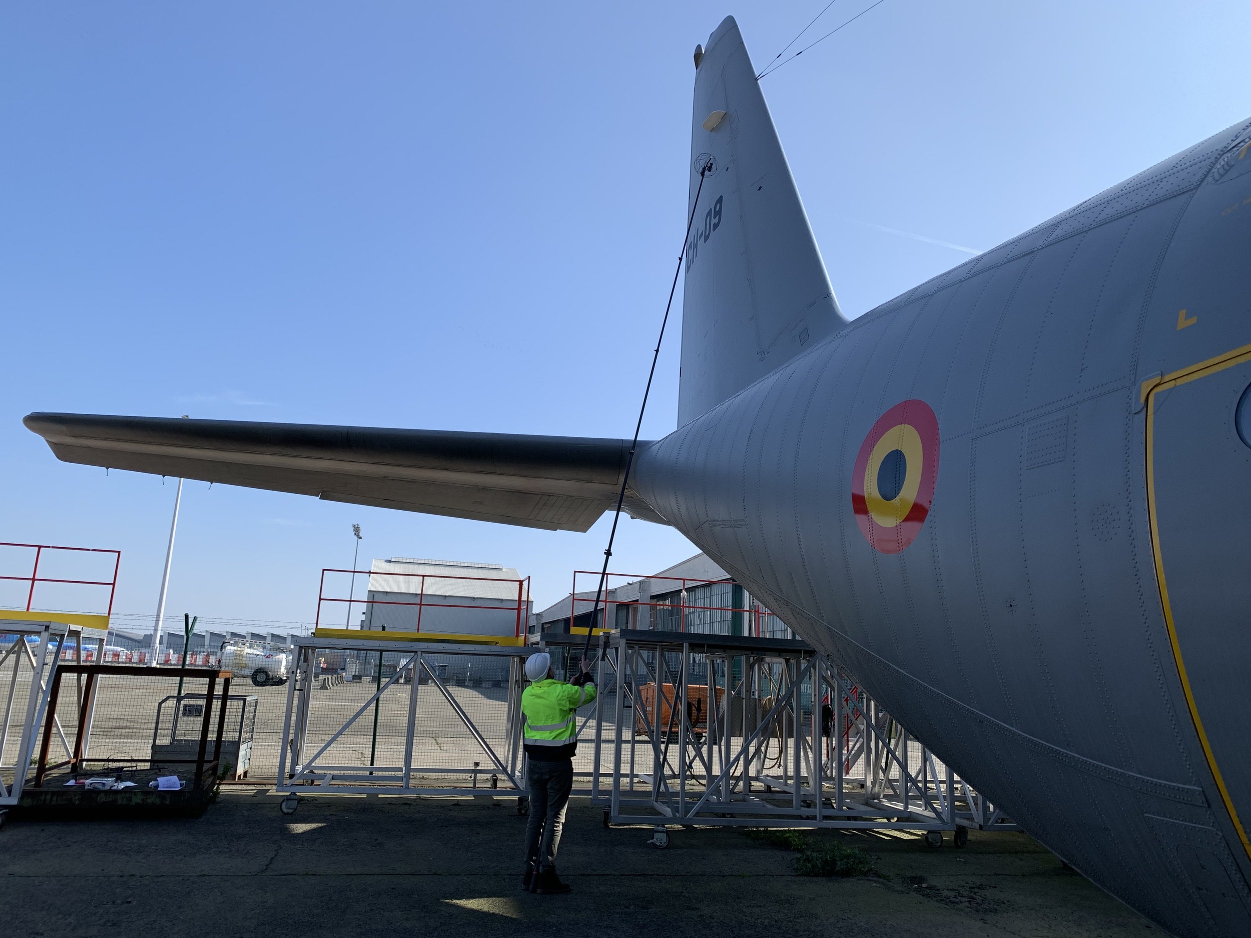 A person in a bright yellow-green jacket and white hard hat is standing underneath the tail section of a military aircraft on a scaffolding platform, with another person nearby, in an outdoor setting with a clear blue sky.