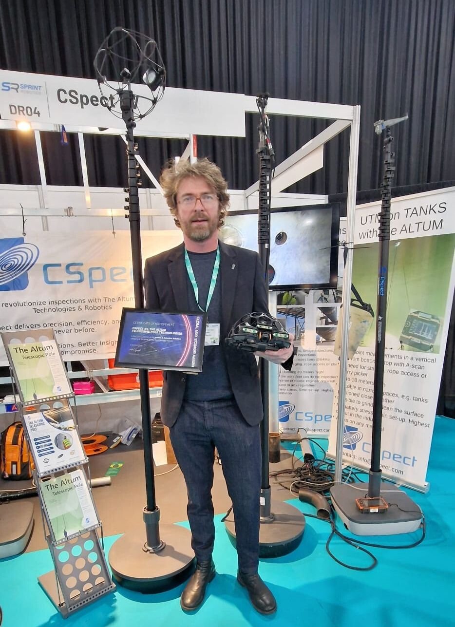 A man with glasses, a beard, and curly hair standing in front of a display at a technology booth, holding a remote control device in his right hand and a tablet in his left. The booth features tall antennas and informational banners about The Altum telescopic pole and inspection technology, with various equipment and banners visible.