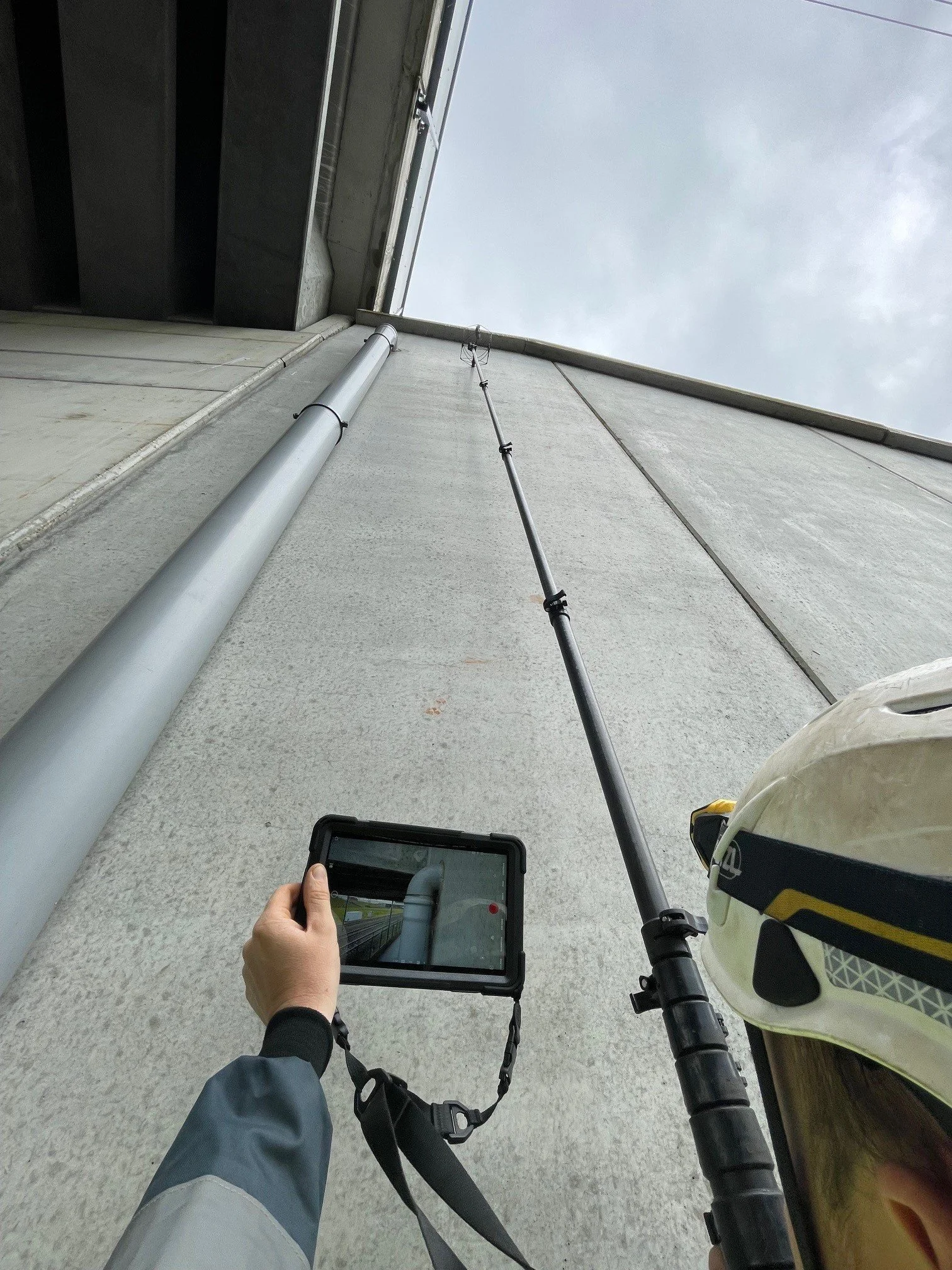 A construction worker's hand is holding a tablet, looking up at an exterior building wall with a long pipe attached to the wall and a safety helmet resting on a bicycle handlebar, all under a cloudy sky.
