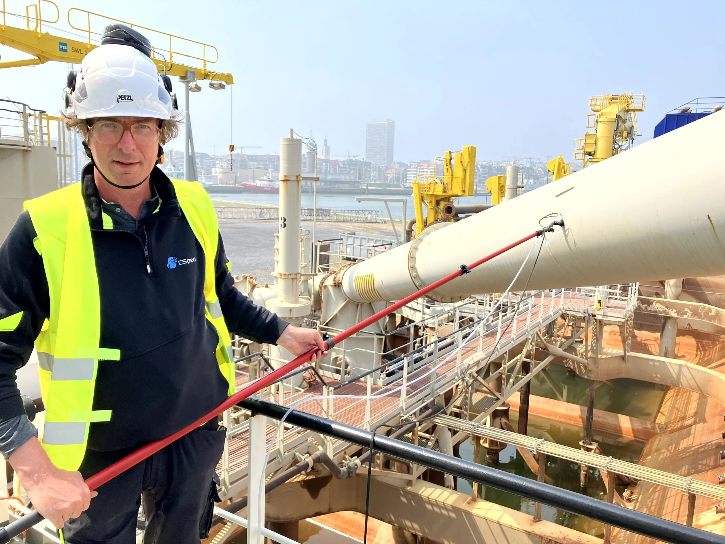 A man wearing safety gear, including a white hard hat and a bright yellow safety vest, stands outdoors near a large industrial pipeline at an industrial site with water, city skyline, and cranes in the background.