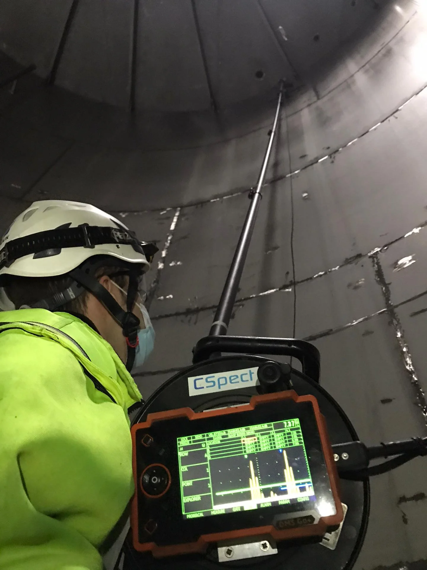 A person wearing a helmet, mask, and high-visibility jacket is using a handheld device with a screen displaying a numerical and graphical interface, inside a metallic, cylindrical structure with visible welding or rivets.