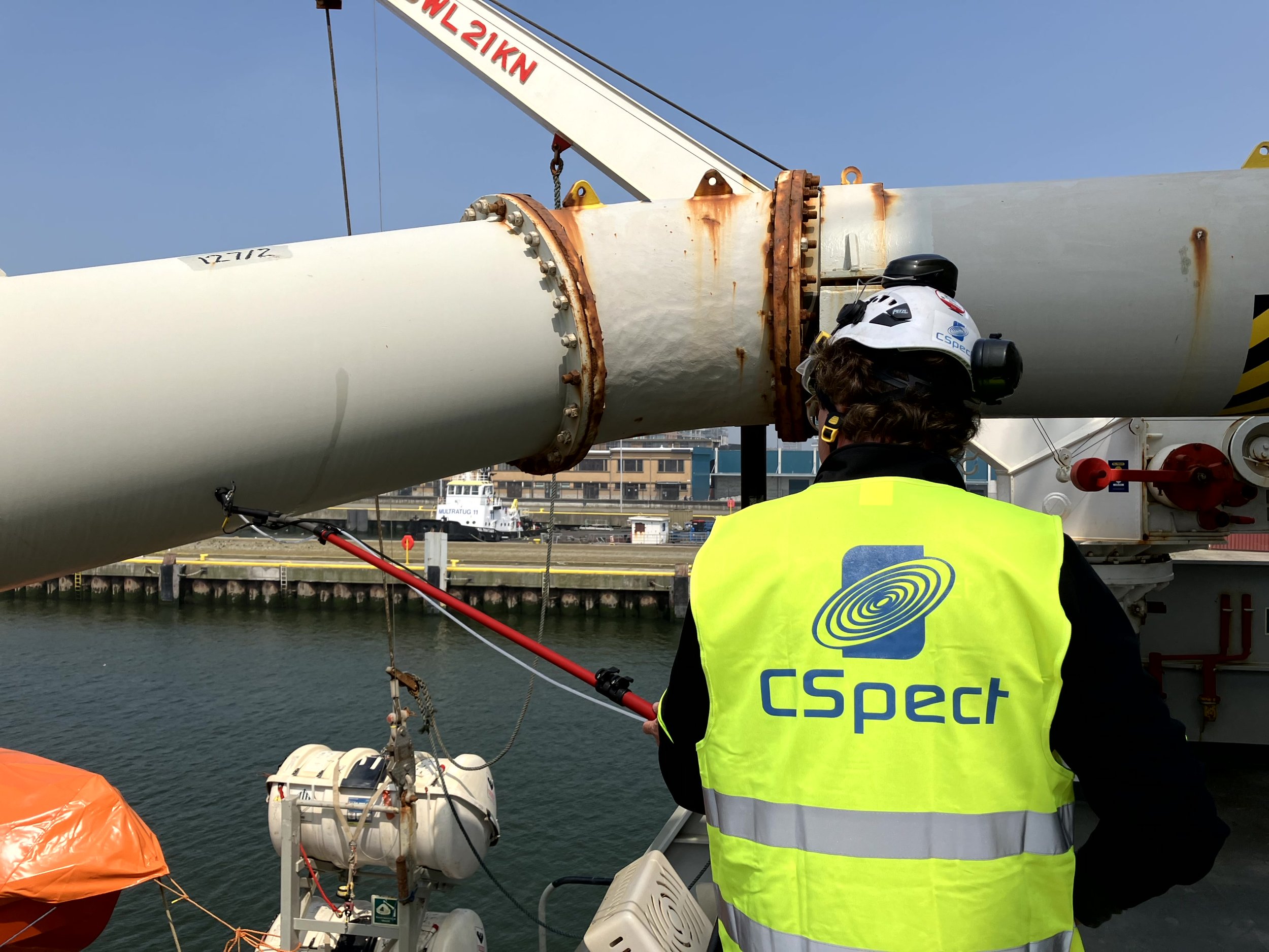 Person wearing a high-visibility vest with 'CSpect' logo inspecting a large industrial pipe or structure near water, with a helmet and safety gear, during daytime.