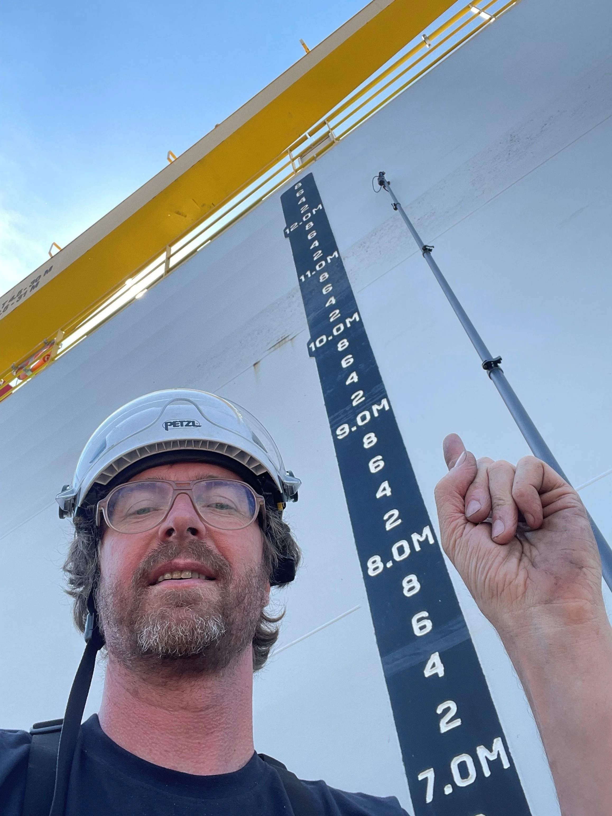 A man wearing a helmet and glasses taking a selfie in front of a large offshore oil or gas platform's side with an height measurement scale from 7 meters to 27 meters.