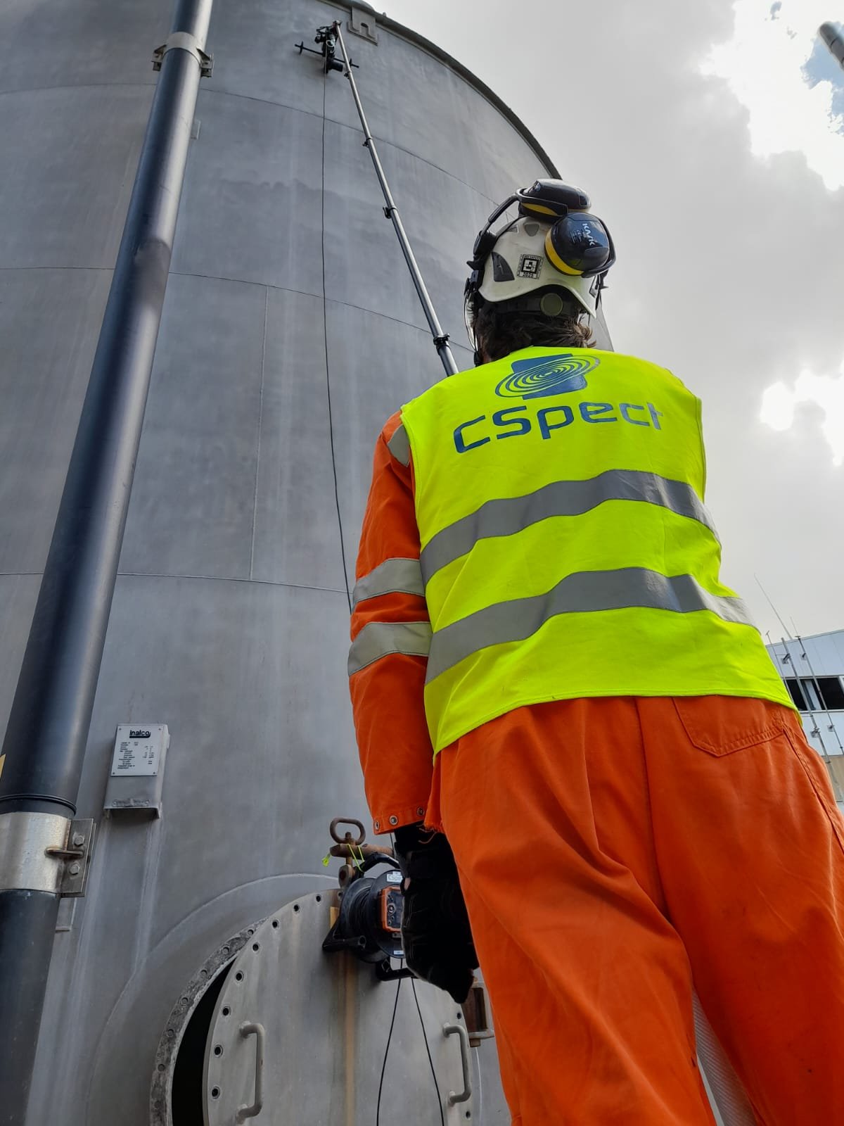 A worker in a high-visibility vest and orange safety pants using a motorized tool to inspect or maintain a large industrial tank, with a safety helmet and hearing protection, against a cloudy sky.