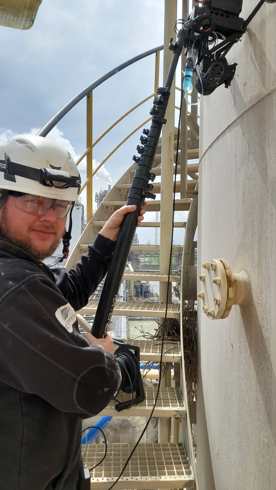 A worker wearing a safety helmet and protective glasses holding a long pipe or rod next to a tall industrial structure outdoors.
