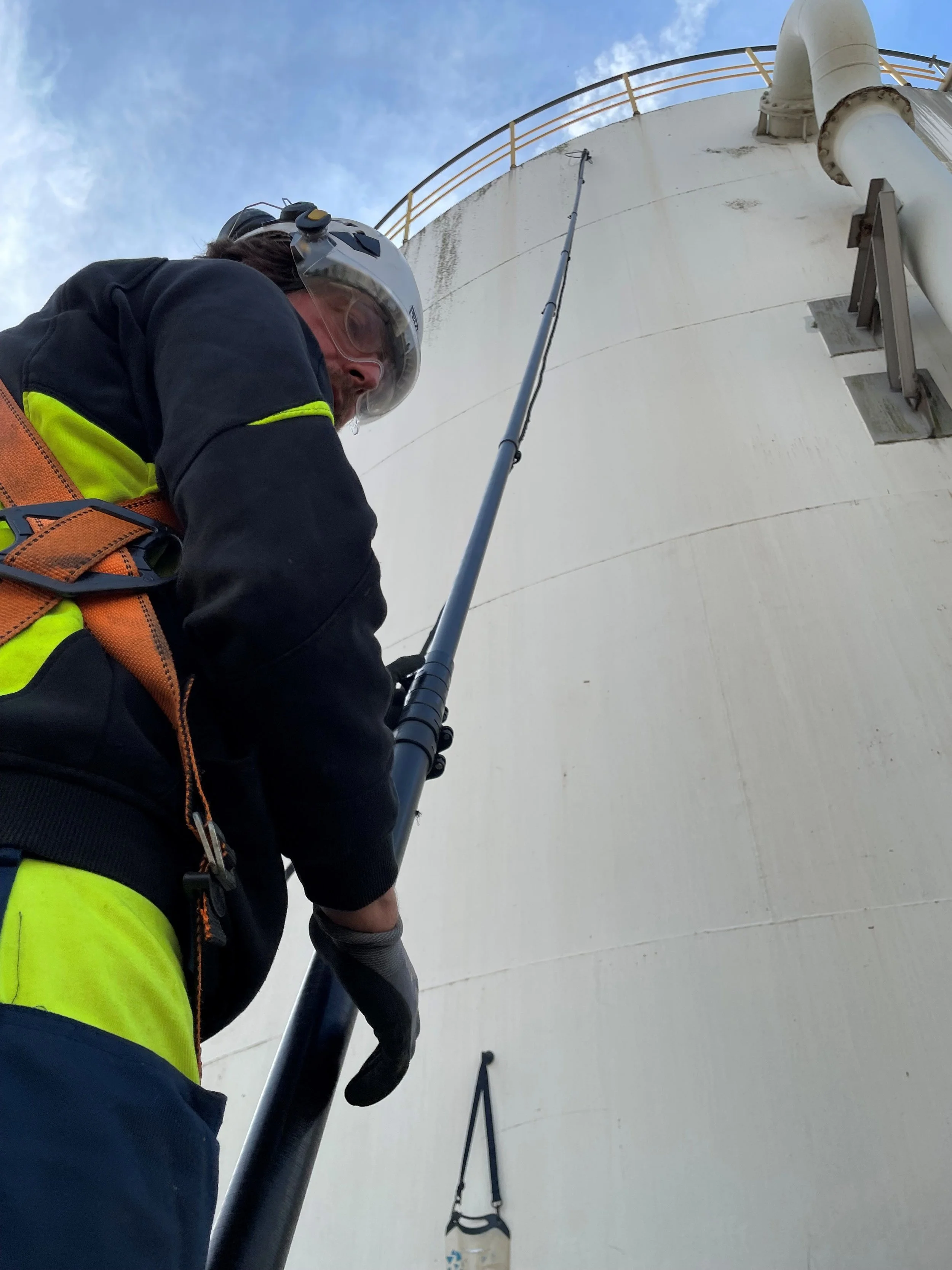 Worker wearing a helmet and safety vest climbing a large industrial tank using a harness and safety rope.