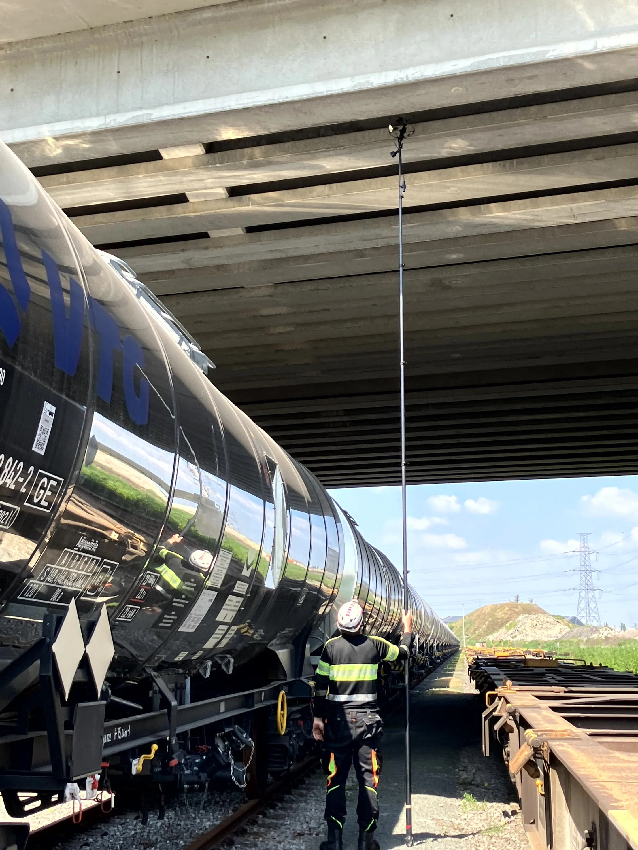 A railway worker in a neon safety vest and helmet looks at a row of shiny black railcars parked under a large concrete bridge. A long measuring pole is positioned beside the worker. The landscape in the background includes grassy hills and transmission towers.