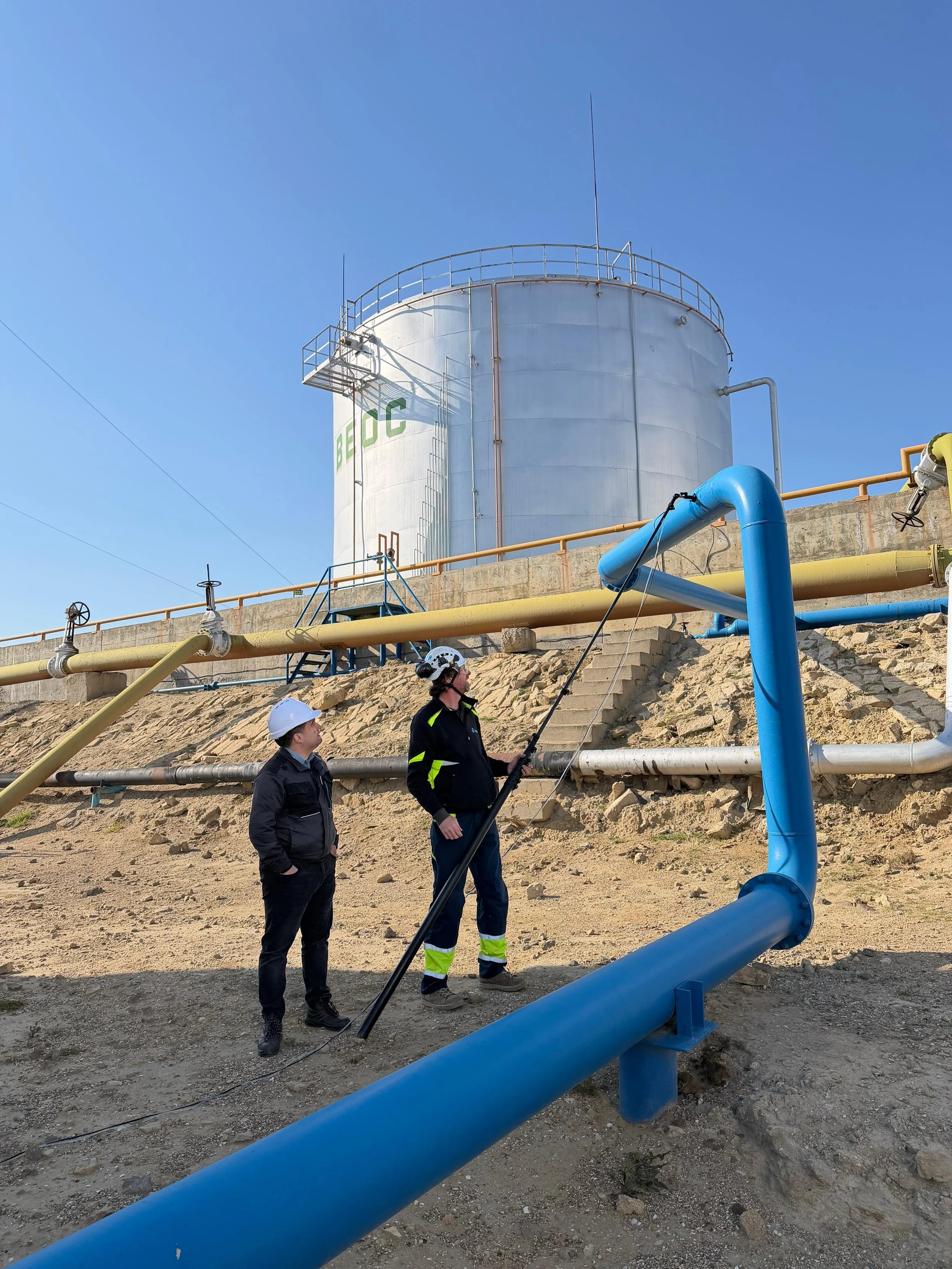 Two workers inspecting industrial pipes outdoors, with a large white storage tank labeled "BEC" and a clear blue sky in the background.
