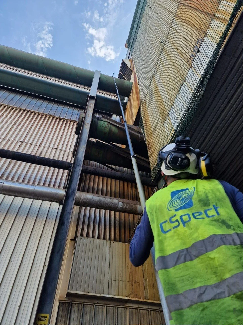 Worker wearing a high-visibility vest with 'CSpect' logo, using a lift or pole to access industrial pipes on a building exterior, with a partly cloudy sky above.