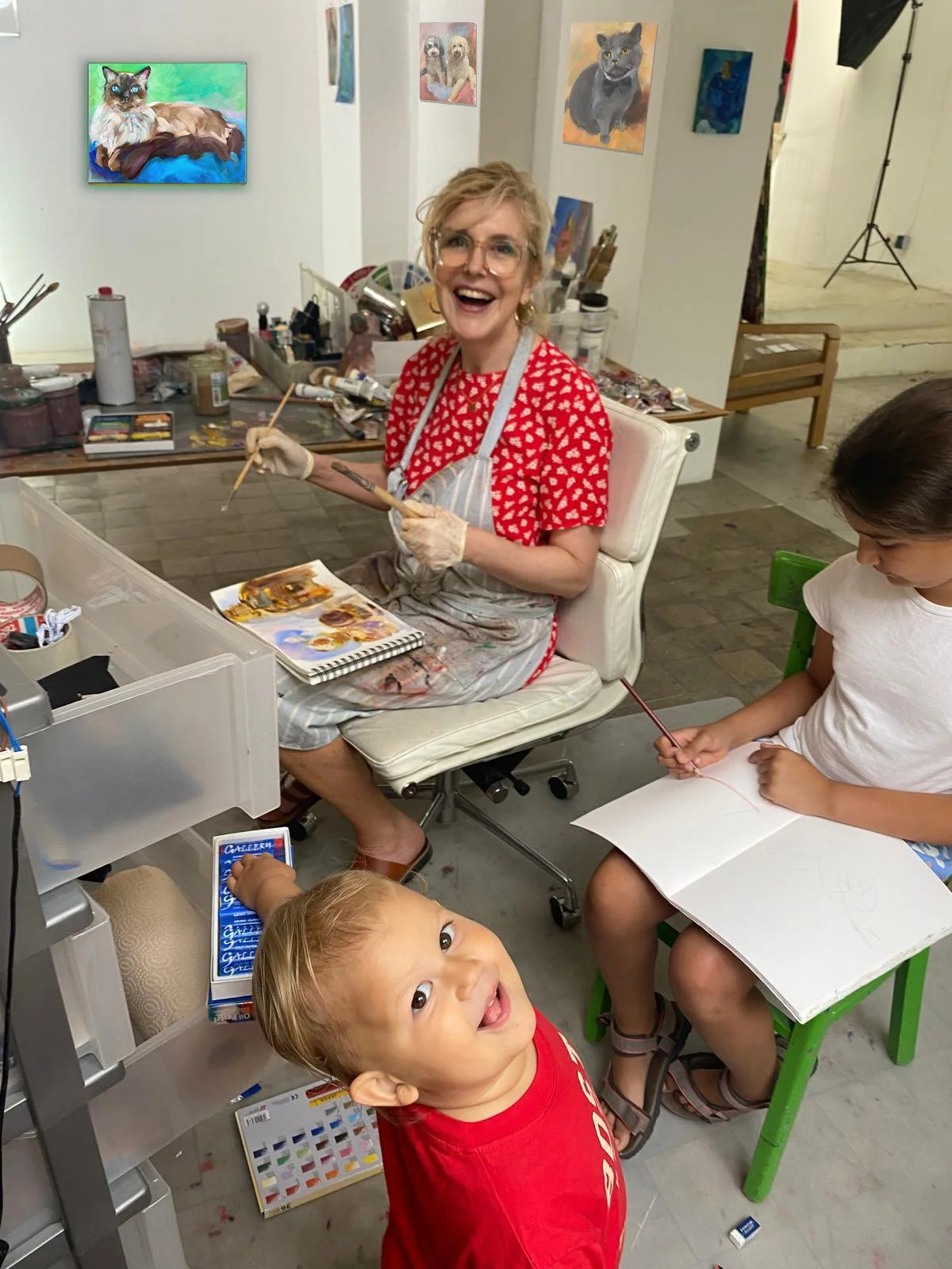 Eine Frau mit Gläsern, vermutlich eine Künstlerin, sitzt an einem Arbeitsplatz in einem Kunststudio, umgeben von Kinder. Sie trägt ein rotes Oberteil mit weißen Herzmustern, eine Schürze und Handschuhe, und hält einen Malpinsel. Zwei Kinder, ein Junge im Vordergrund mit einem roten T-Shirt, schaut lächelnd in die Kamera, während ein anderes Mädchen auf der rechten Seite mit einem Pinsel in der Hand zeichnet. Im Hintergrund sind mehrere Gemälde von Tieren an den Wänden, darunter Katzen und Hunde.