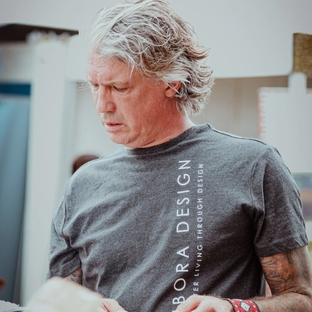 A man with gray hair looking down, wearing a gray t-shirt with white text that reads 'ABORA DESIGN' and 'NEVER LIVING THROUGH DESIGN'.