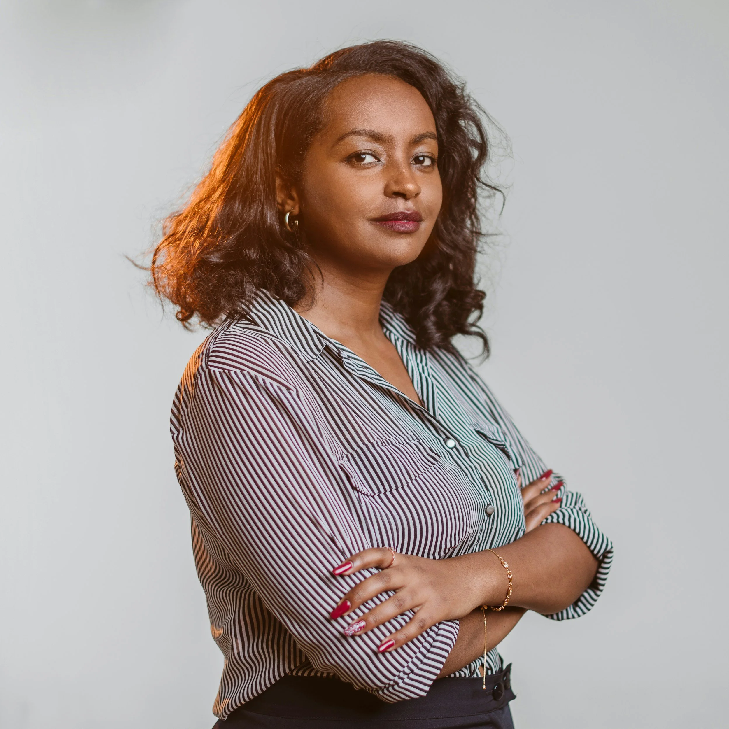 A woman with shoulder-length curly hair wearing a striped button-up shirt with her arms crossed, against a plain light background.