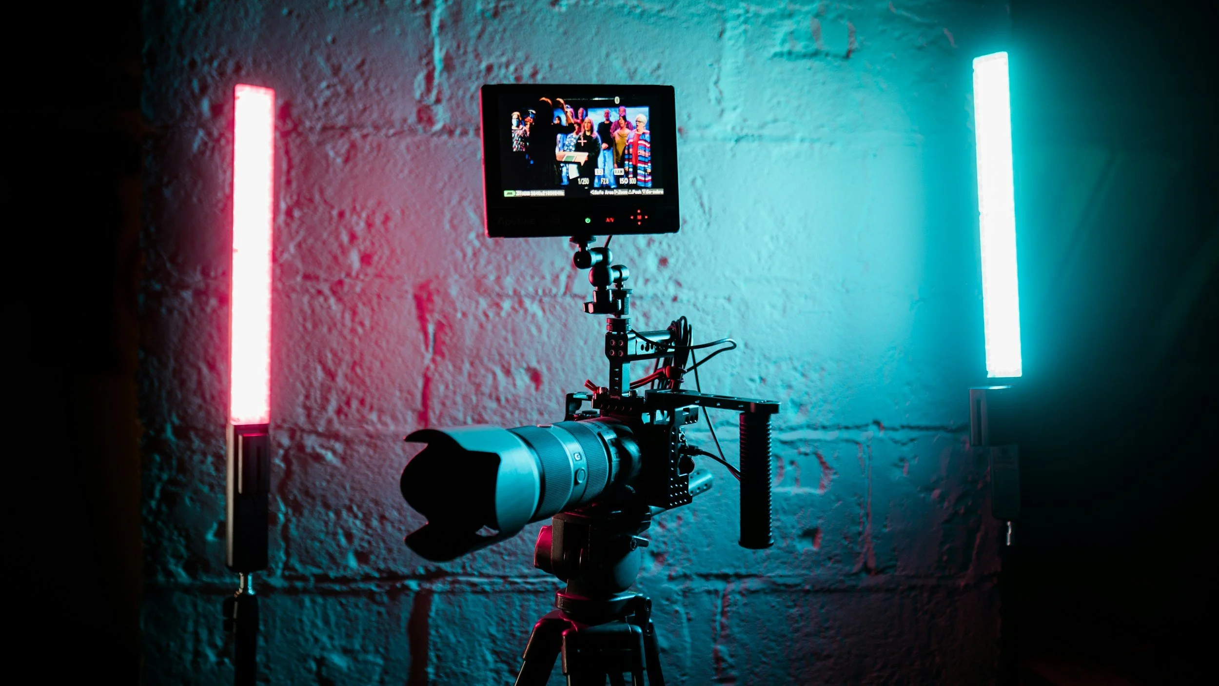 Camera setup with a monitor on a tripod, surrounded by blue and pink LED lights, against a textured white wall