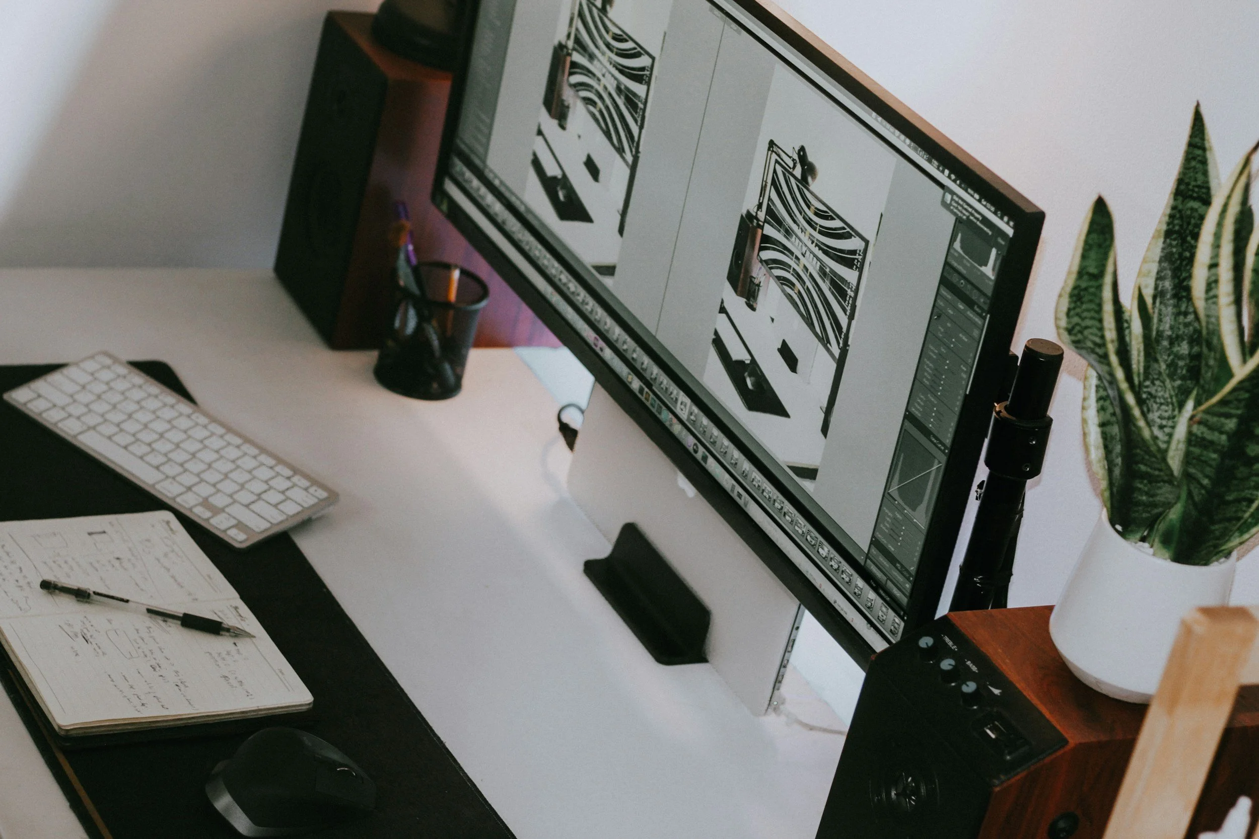 A desk setup with a large computer monitor displaying graphic design work, a wireless keyboard, a mouse on a black desk mat, a notebook with handwritten notes and a pen, a small black container holding pens, and a potted snake plant on a wooden stand.