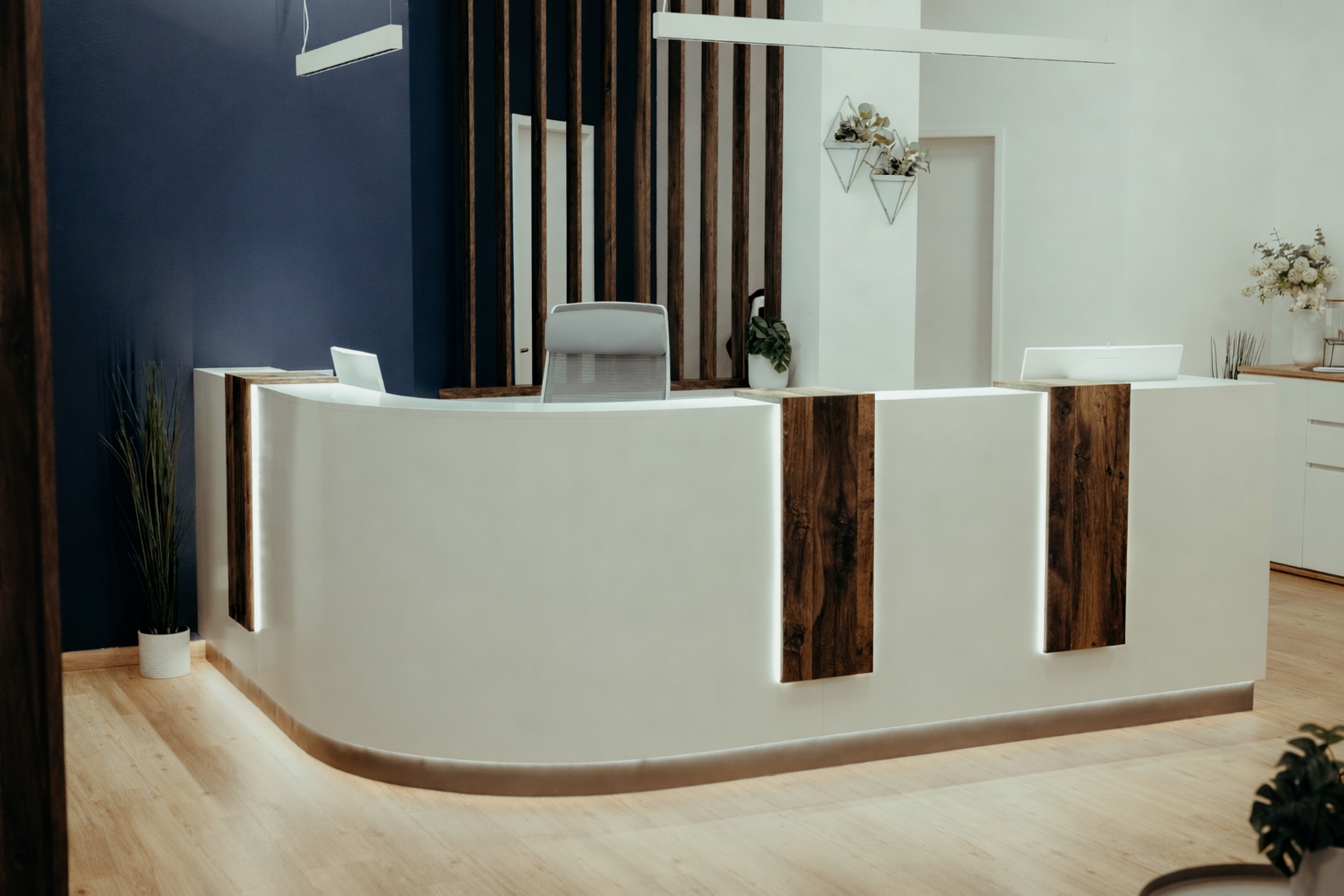 Moderne Empfangstresen im Wartebereich mit weißen Fronten, dunklen Holzleisten und Pflanzen, vor einer dunklen blauen Wand und weißen Wänden mit Dekorationen.