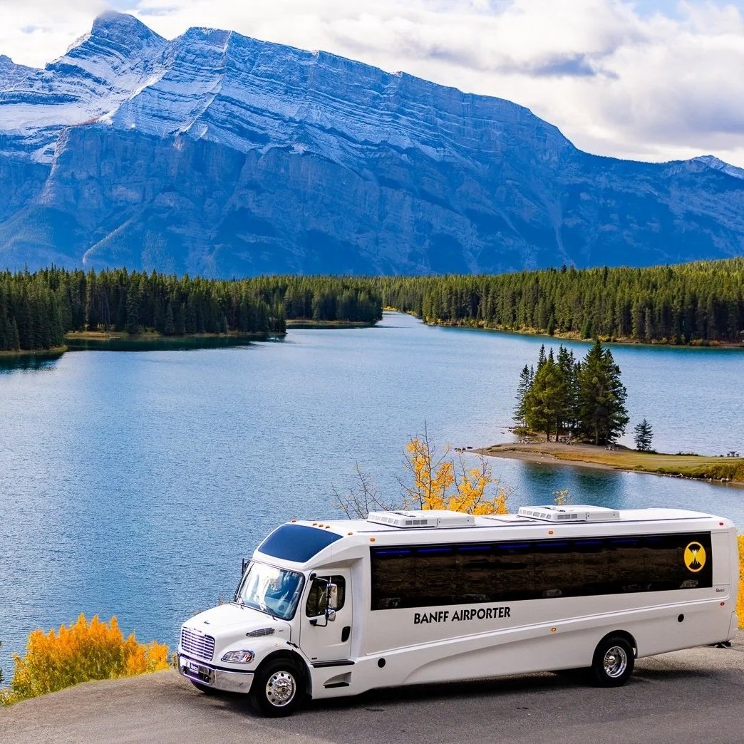 A white bus labeled 'Banff Airporter' parked on a roadside with a scenic lake, dense forest, and tall mountain in the background.
