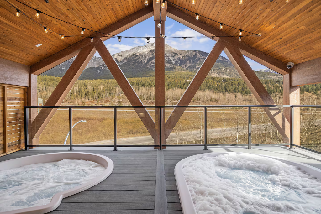 Two hot tubs on a balcony with a mountain view in the background, surrounded by a forest and under a wooden ceiling with string lights.