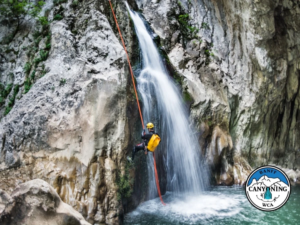 A person rappelling down a waterfall on a steep rocky canyon wall, wearing a helmet, harness, and yellow backpack. The logo of Banff Canyoning is in the lower right corner.