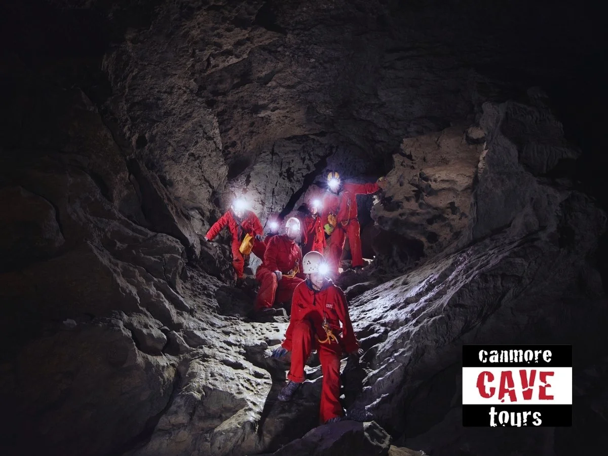 Group of people in red protective suits and helmets exploring a dark cave with headlamps on. The bottom right corner has a logo that reads 'Cannmore Cave Tours'.