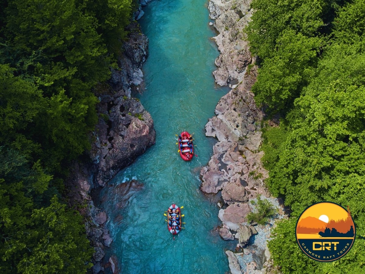 Two red inflatable rafts with people white water rafting in a narrow river surrounded by rocky banks and dense green trees, with a logo of Camdmore Raft Tours in the bottom right corner.