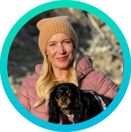 A woman wearing a tan knit hat and a pink puffer jacket smiling outdoors with a small black and brown dog.