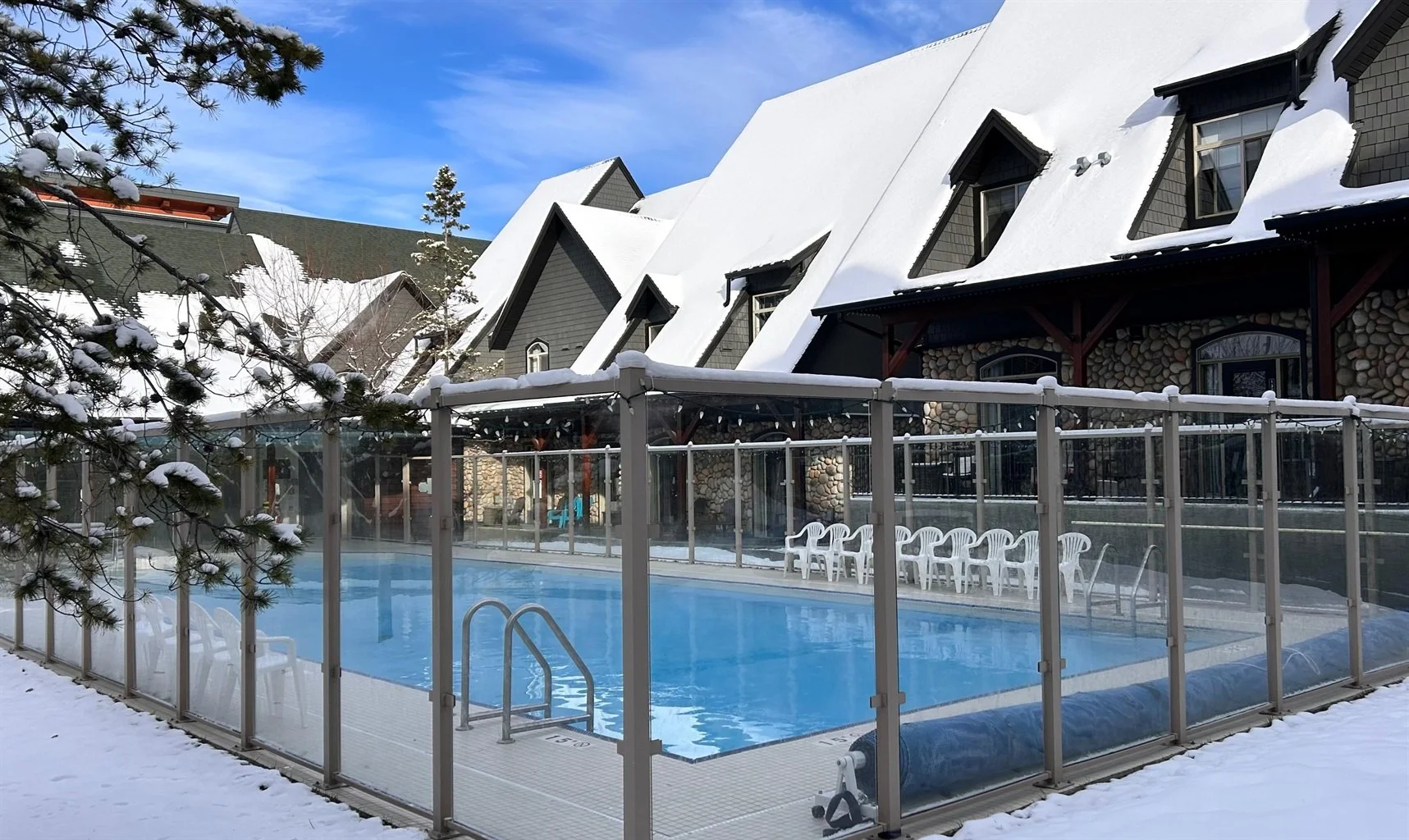 A snow-covered outdoor swimming pool with a glass enclosure, surrounded by snow, in front of houses with snow on their roofs, during daytime with a partly cloudy sky.