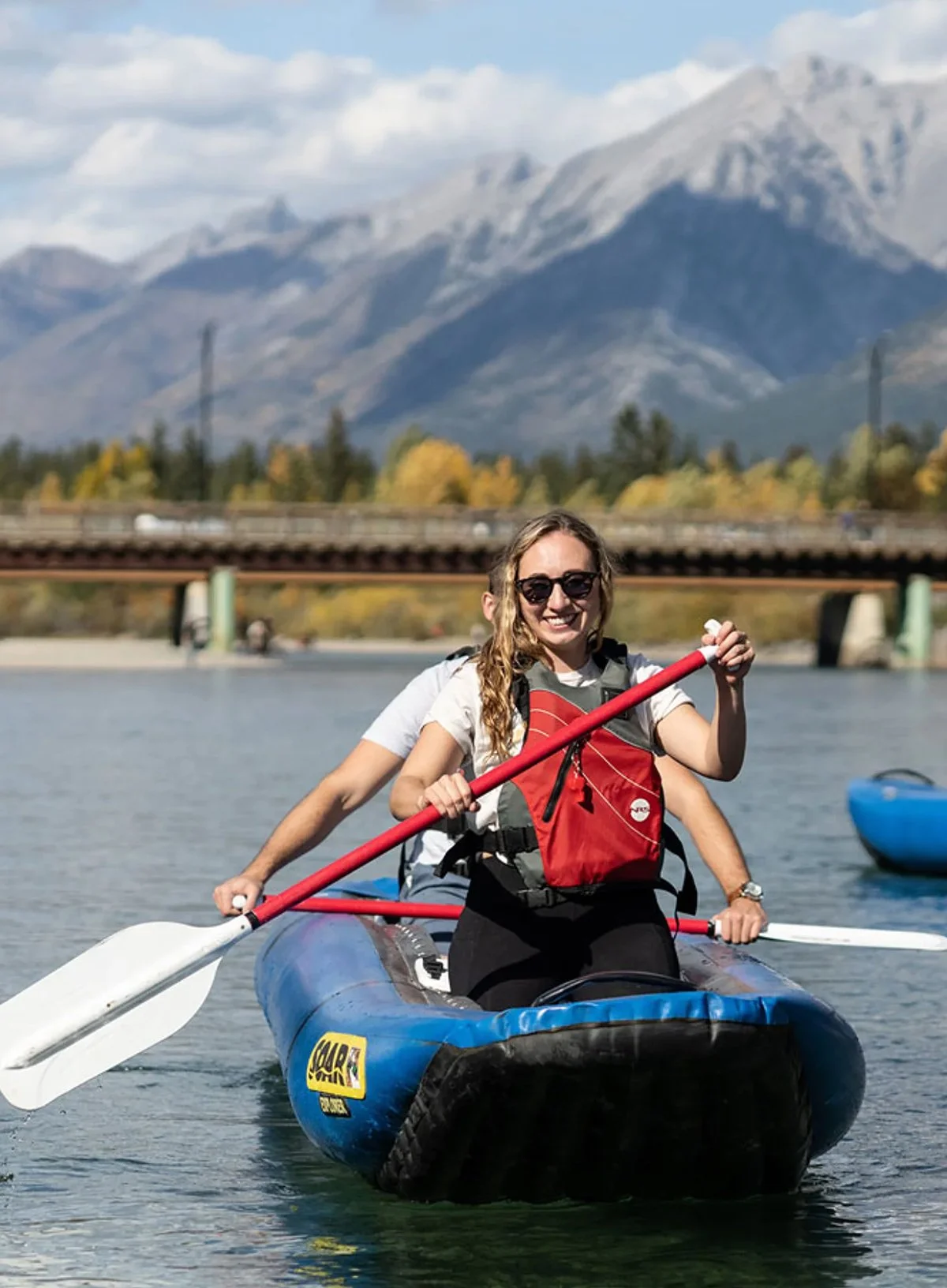 A woman smiling while kayaking on a river with landscapes of mountains and trees in the background.