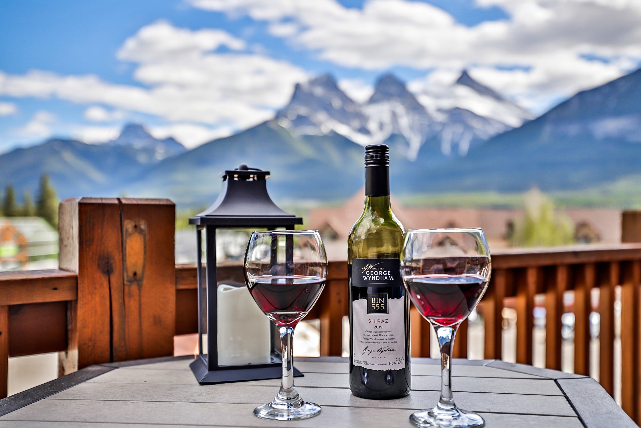 A table set outdoors with two glasses of red wine, a bottle of George Wyndham Shiraz wine, and a candle in a lantern, with a mountain landscape in the background.