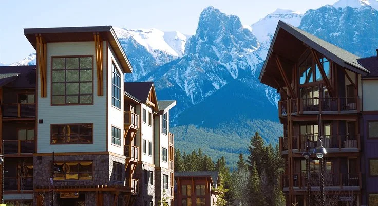 Residences with mountain view, trees in foreground, snow-capped peaks in background.