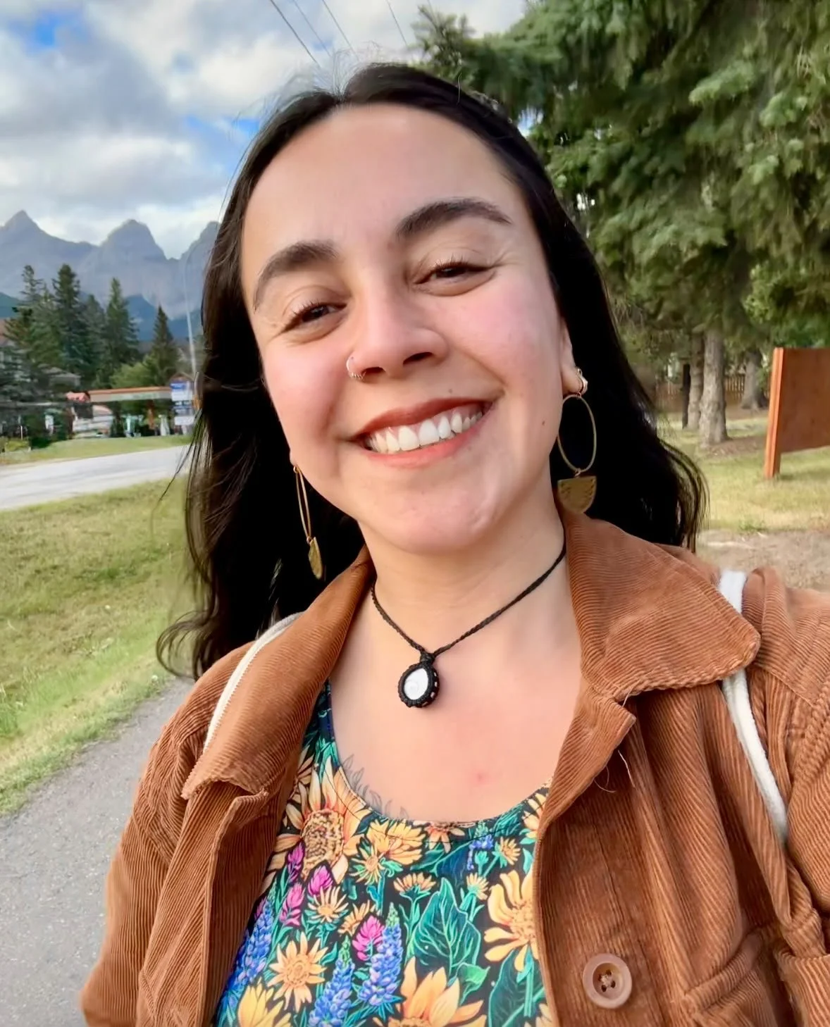 A smiling woman with dark hair, wearing hoop earrings, a black necklace with a pendant, a floral shirt, and a brown jacket, outdoors with trees and mountains in the background.
