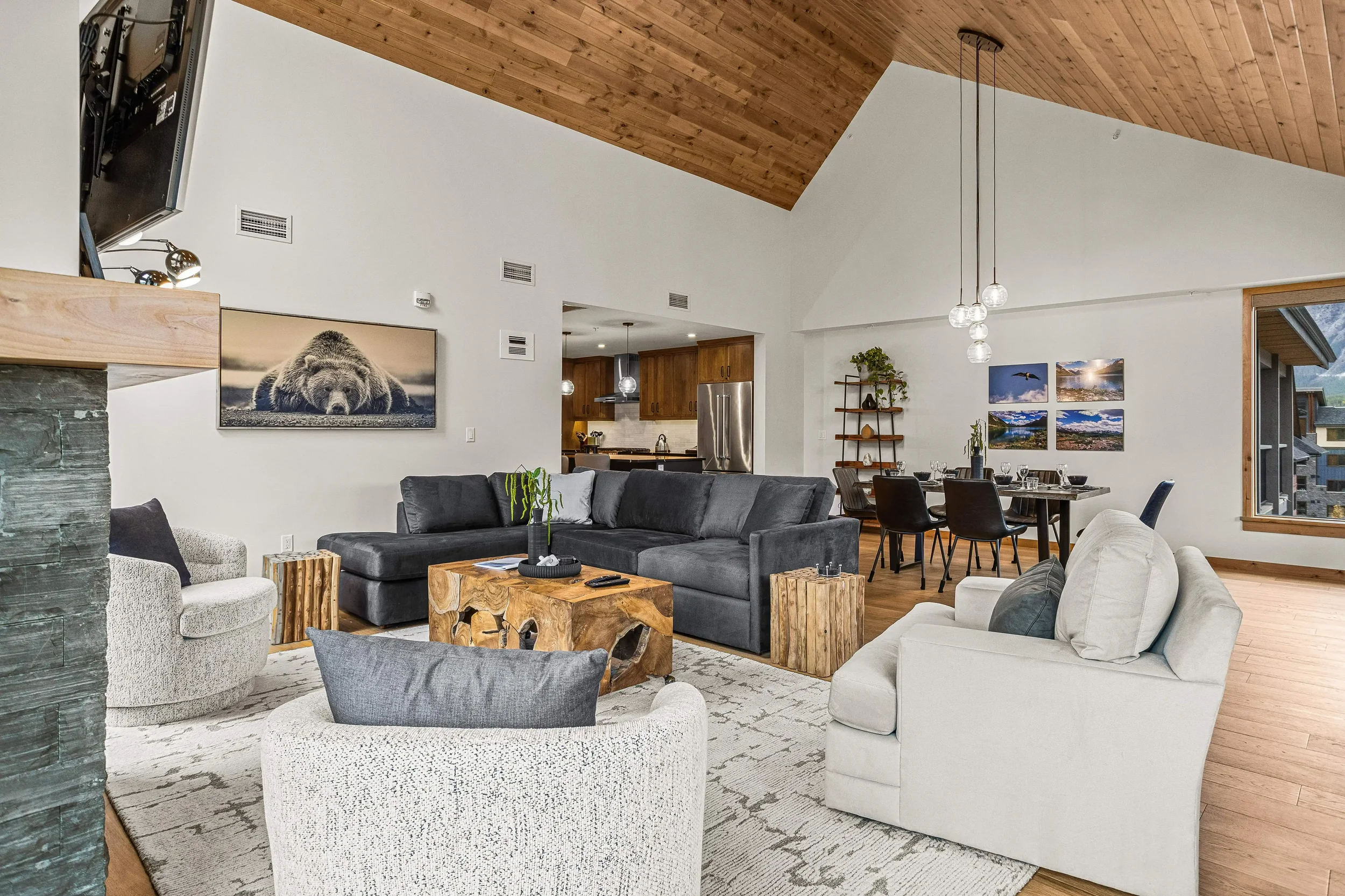 Living room with white walls, vaulted wooden ceiling, gray sectional sofa, white armchair, and wooden side tables. Open kitchen with wooden cabinets and stainless steel appliances in the background. Dining table with black chairs and hanging globe lights. Wall art including a bear print and landscape photos, window with mountain view.