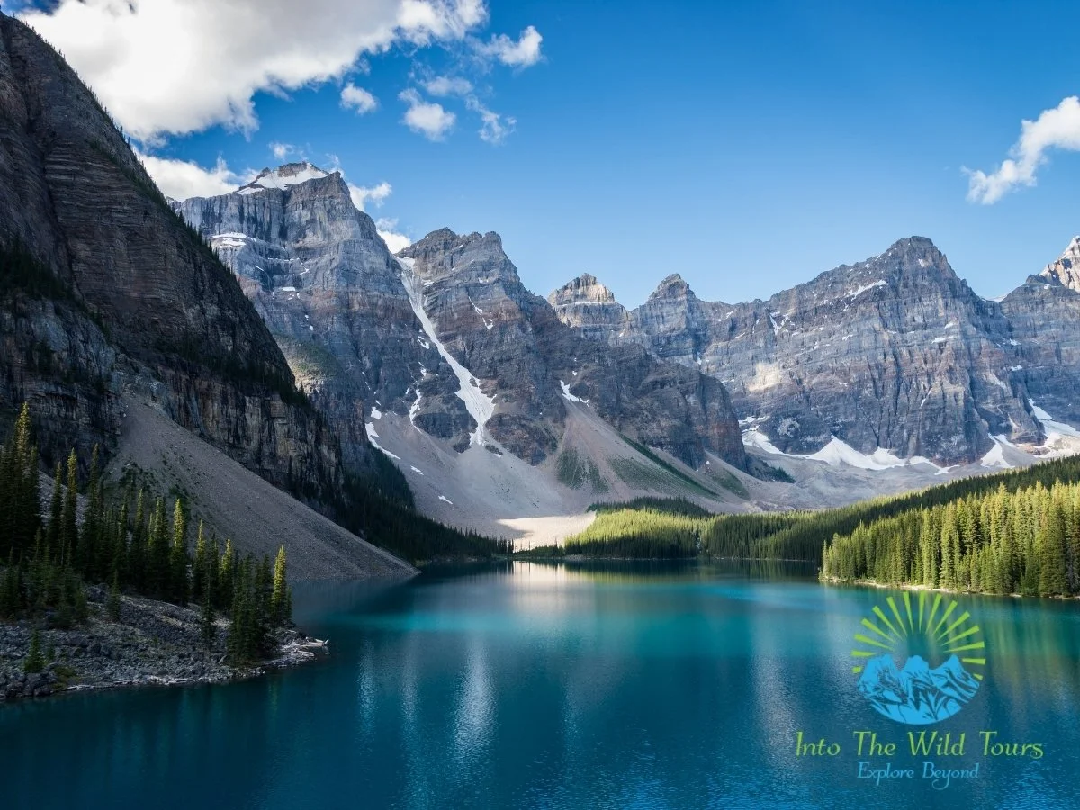 Scenic view of a mountain lake with clear blue water, surrounded by dense green forest, steep rocky mountains with patches of snow, and a partly cloudy sky. Logo of 'Into The Wild Tours' with the tagline 'Explore Beyond' in the bottom right corner.
