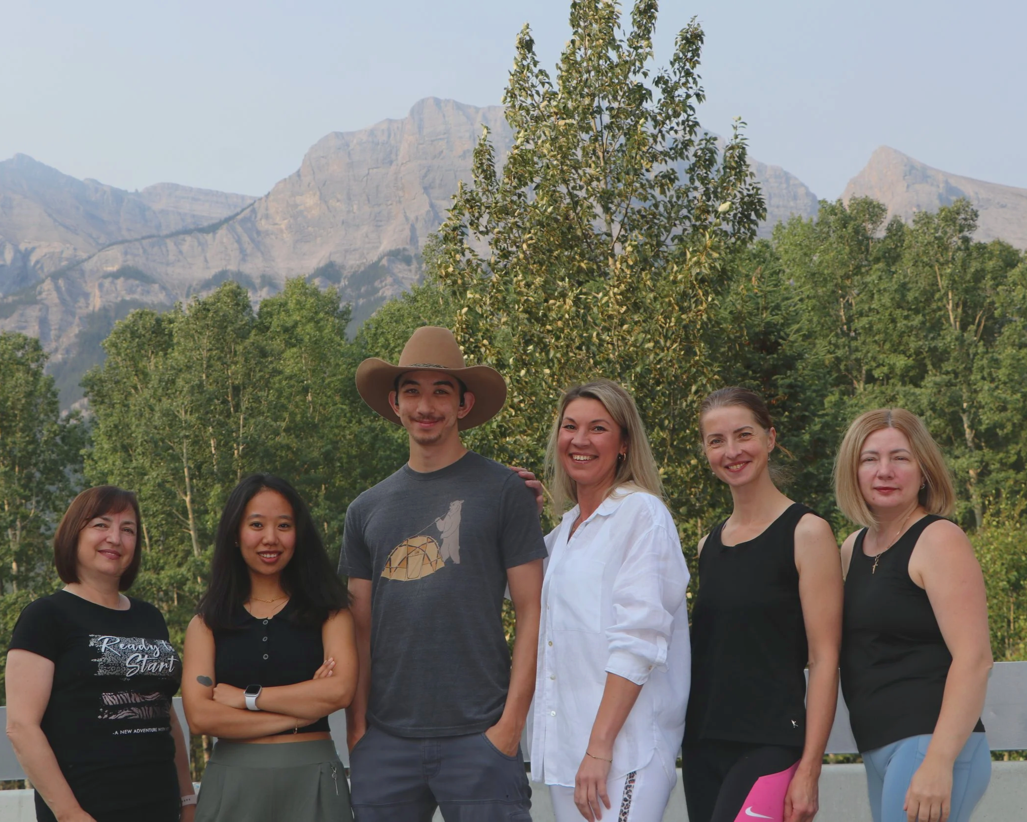 A group of diverse smiling people, mostly women, standing outdoors with mountains and trees in the background.
