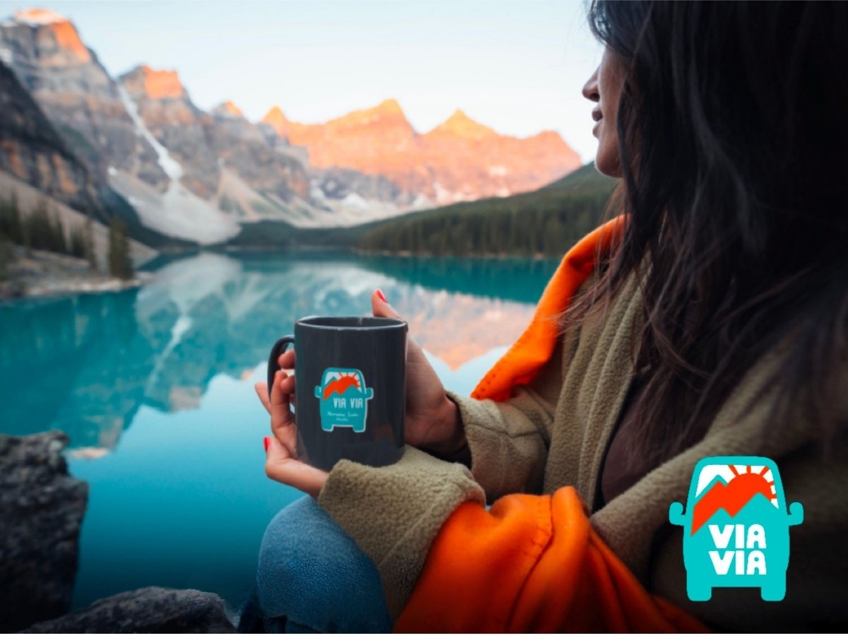 A woman sitting by a lake holding a black mug with a Via Via logo, overlooking mountains and water reflecting the landscape.
