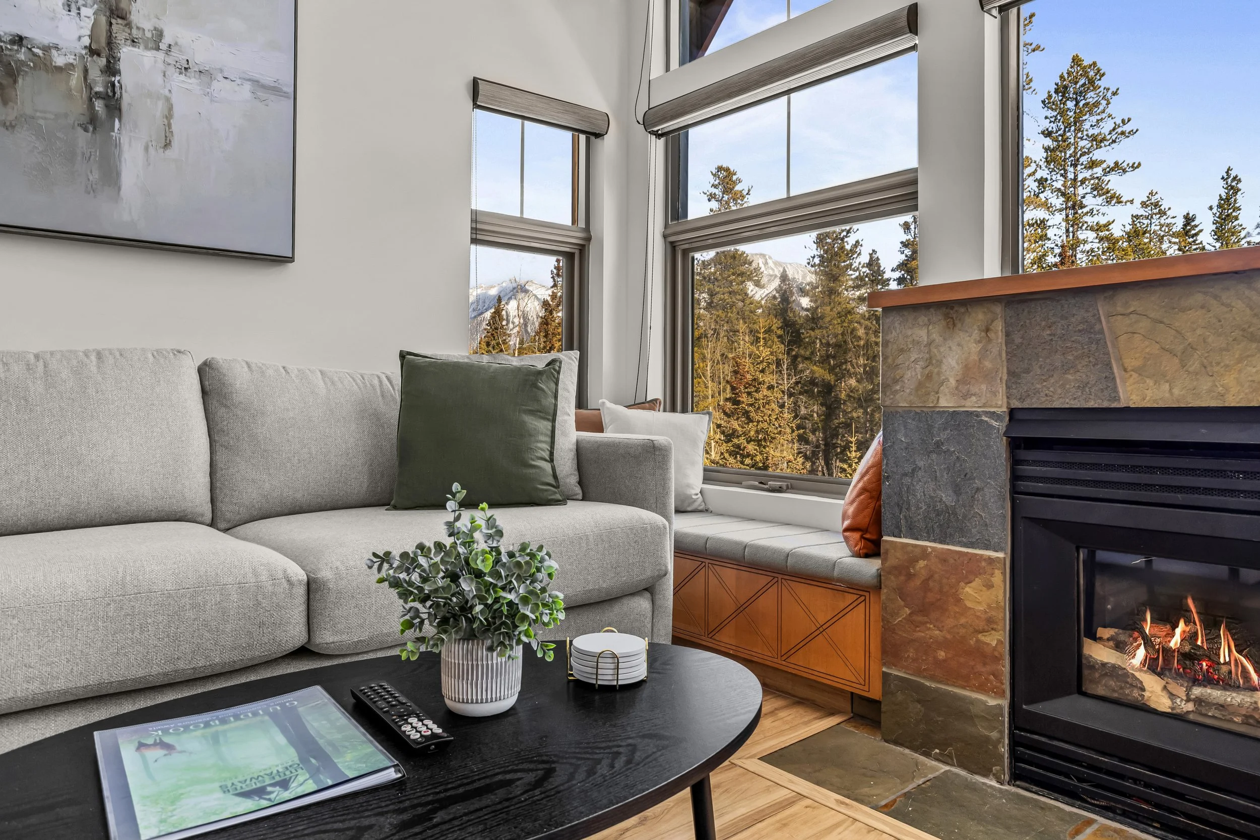 Living room with a gray sofa, green and white pillows, a black coffee table with a plant, remote, and magazine, large windows showing trees and mountains, and a fireplace with a small fire.