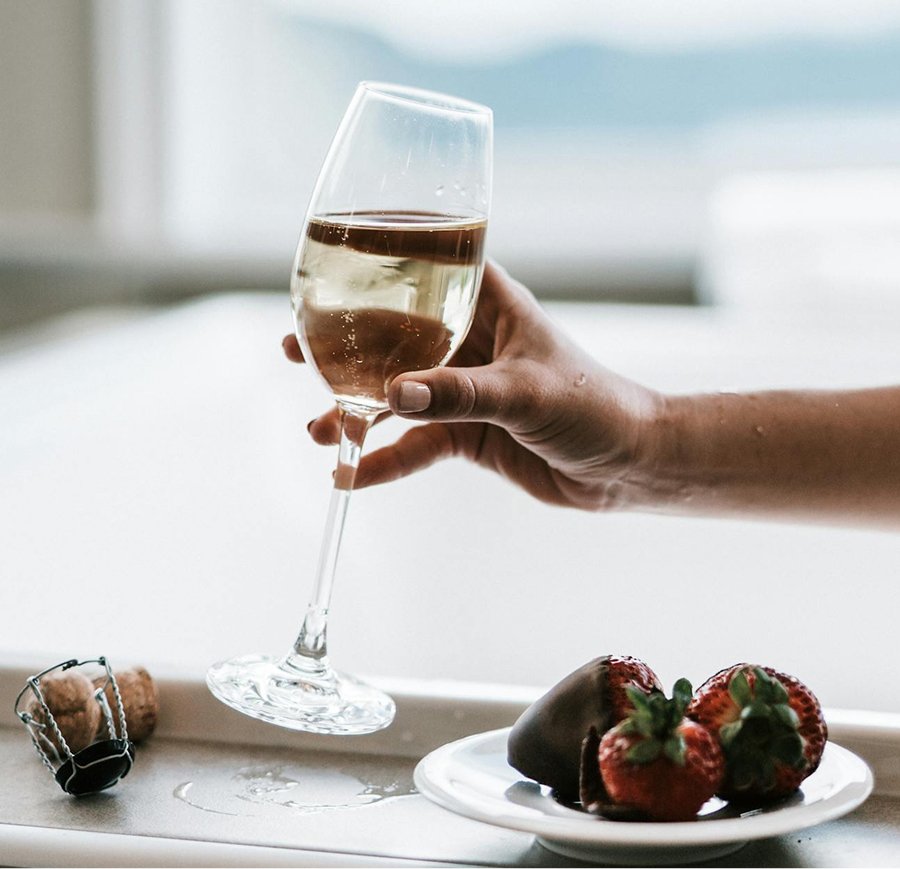 A hand holding a glass of sparkling wine or champagne, with a plate of strawberries and a chocolate-covered strawberry nearby, on a white surface near a window.